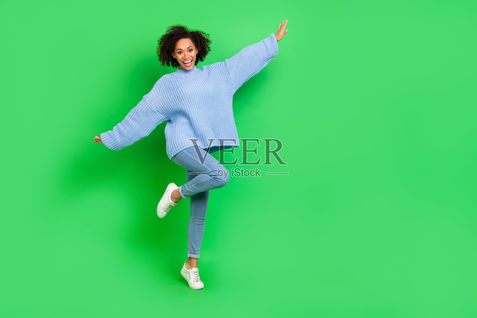 可爱的小女孩全身照,卷发张开双手欢笑,穿着时尚的针织毛衣,背景为绿色。照片摄影图片