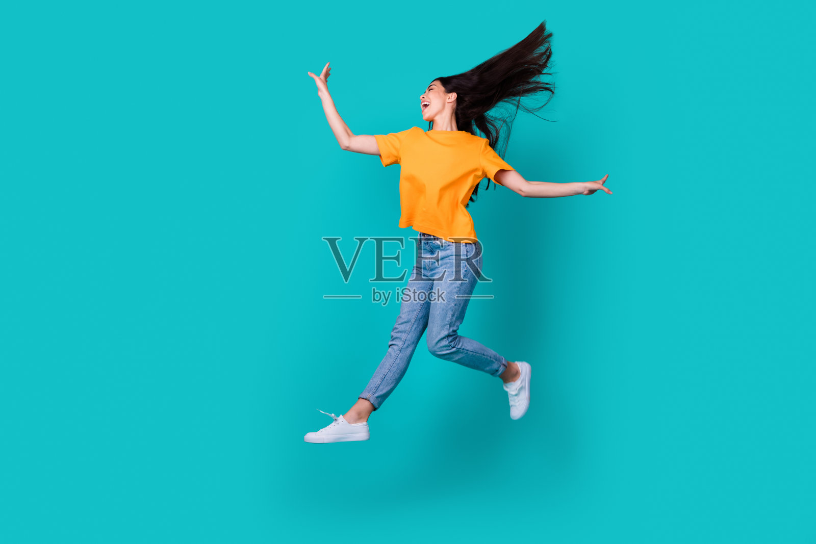 全身照,酷炫的棕发女士穿着橙色T恤、牛仔裤和靴子,背景为青绿色。照片摄影图片