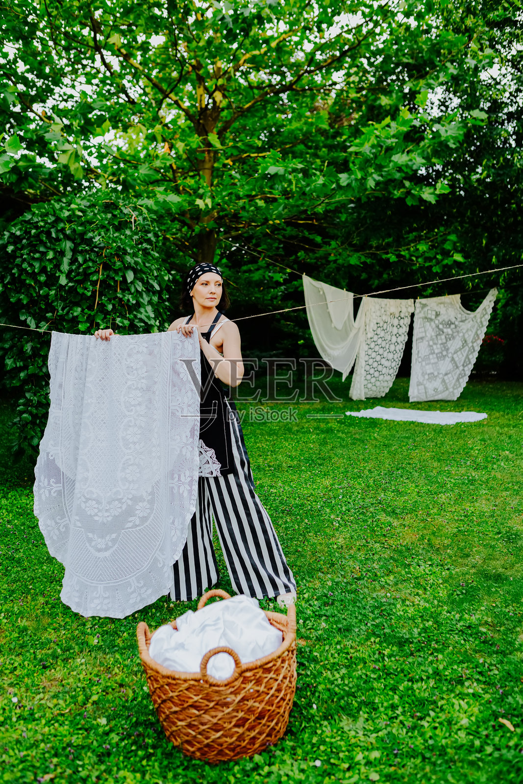 女人在郁郁葱葱的花园里,正在晾晒精美的蕾丝布料。照片摄影图片
