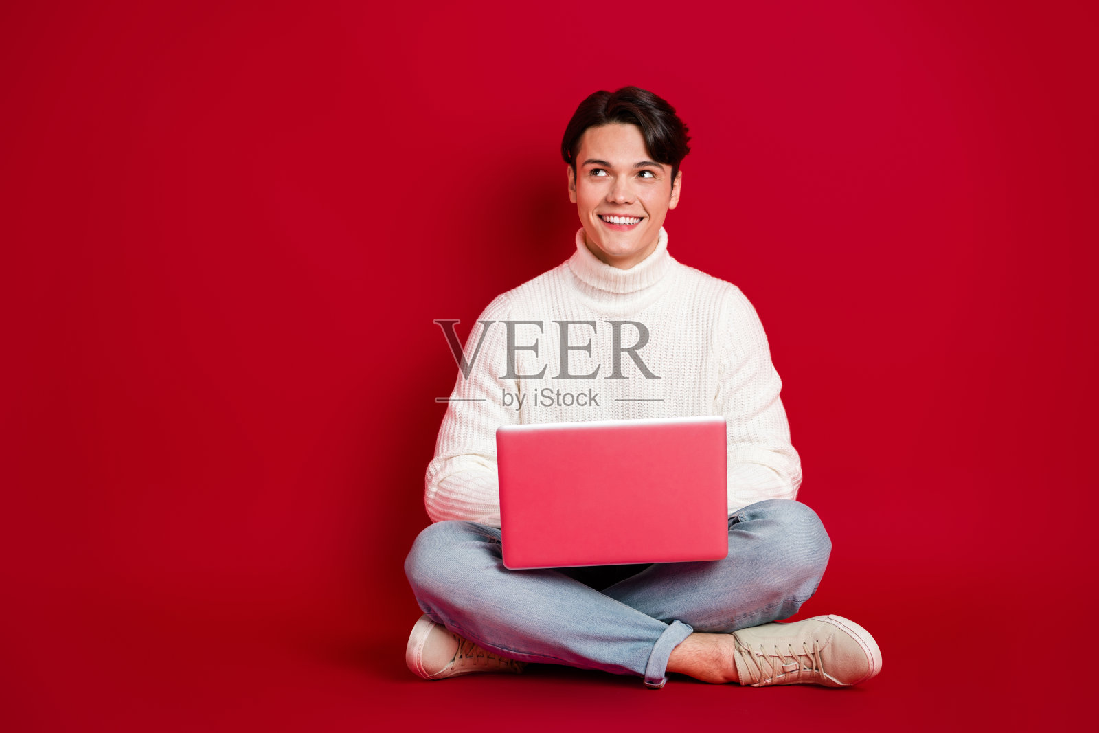 全身照,帅气的男孩坐在地板上,使用笔记本电脑,目光投向空旷的空间,背景为红色。照片摄影图片