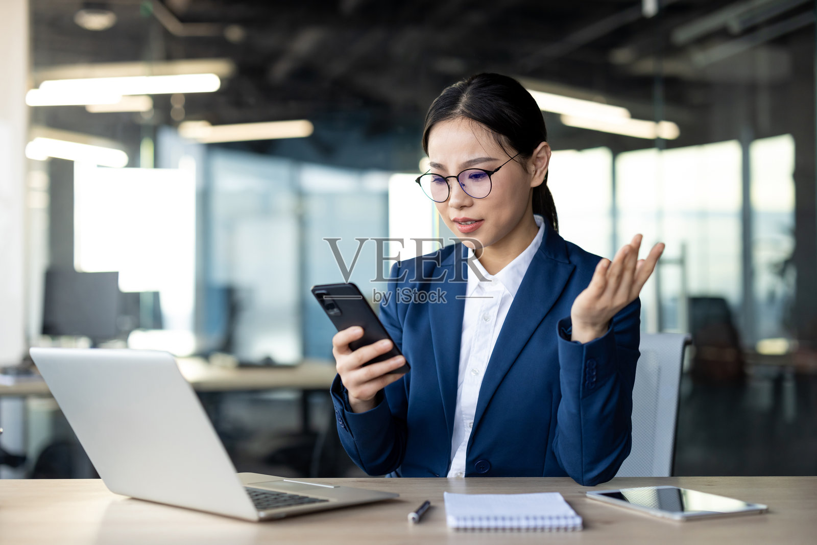 在办公室一边看手机一边用笔记本电脑工作的亚洲女商人,面露困惑或沉思照片摄影图片