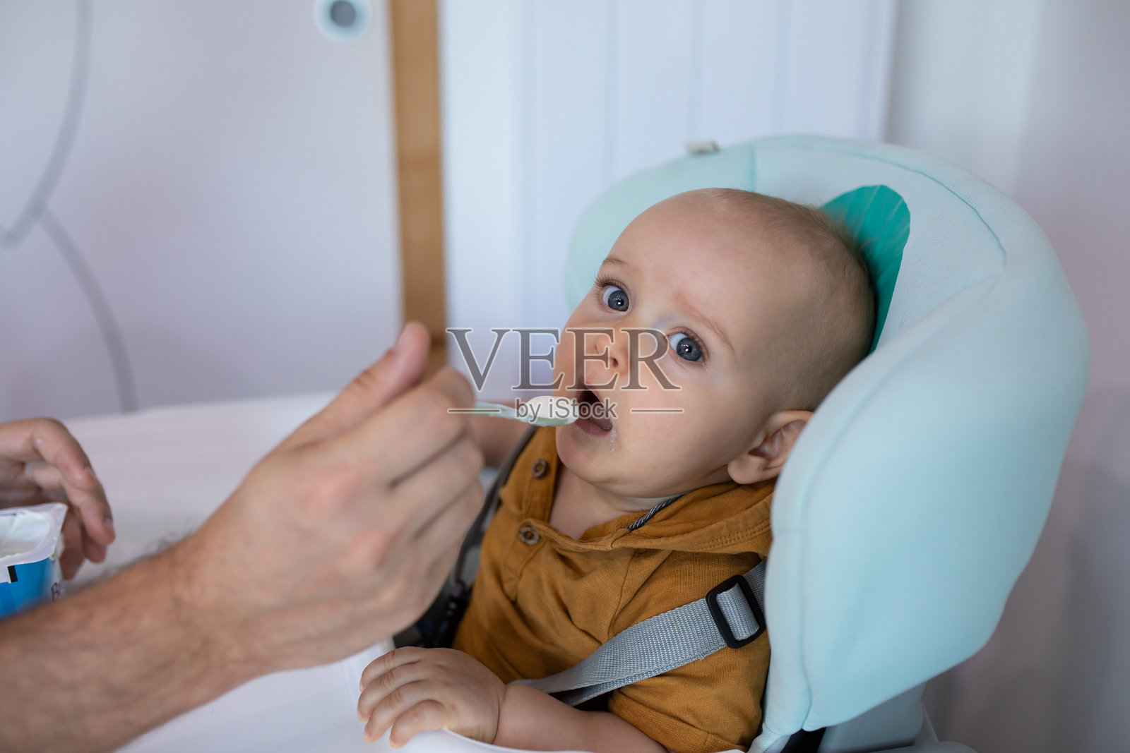 婴儿在高椅上被一只男人的手用勺子喂食,表现出接受、认真,正在吃他的第一口固体食物,进行食物多样化。照片摄影图片
