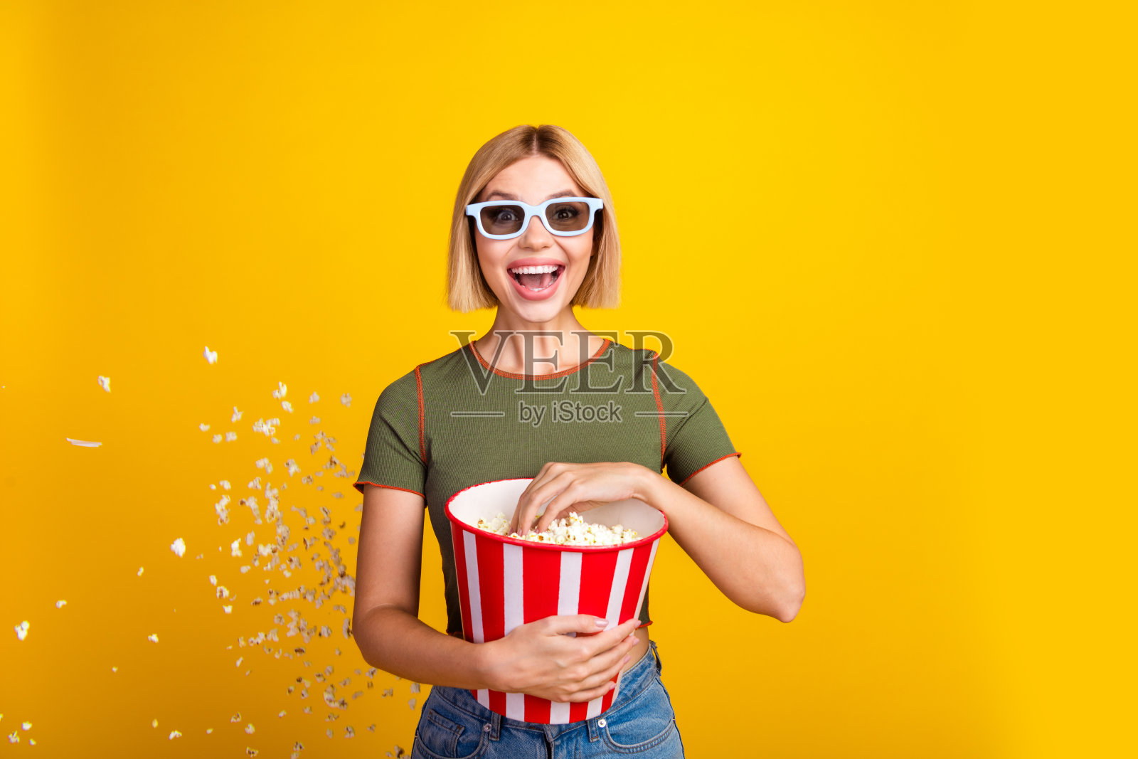 戴着3D眼镜、穿着卡其色T恤的短发女孩,惊讶地吃着爆米花观看首映,背景为黄色。照片摄影图片