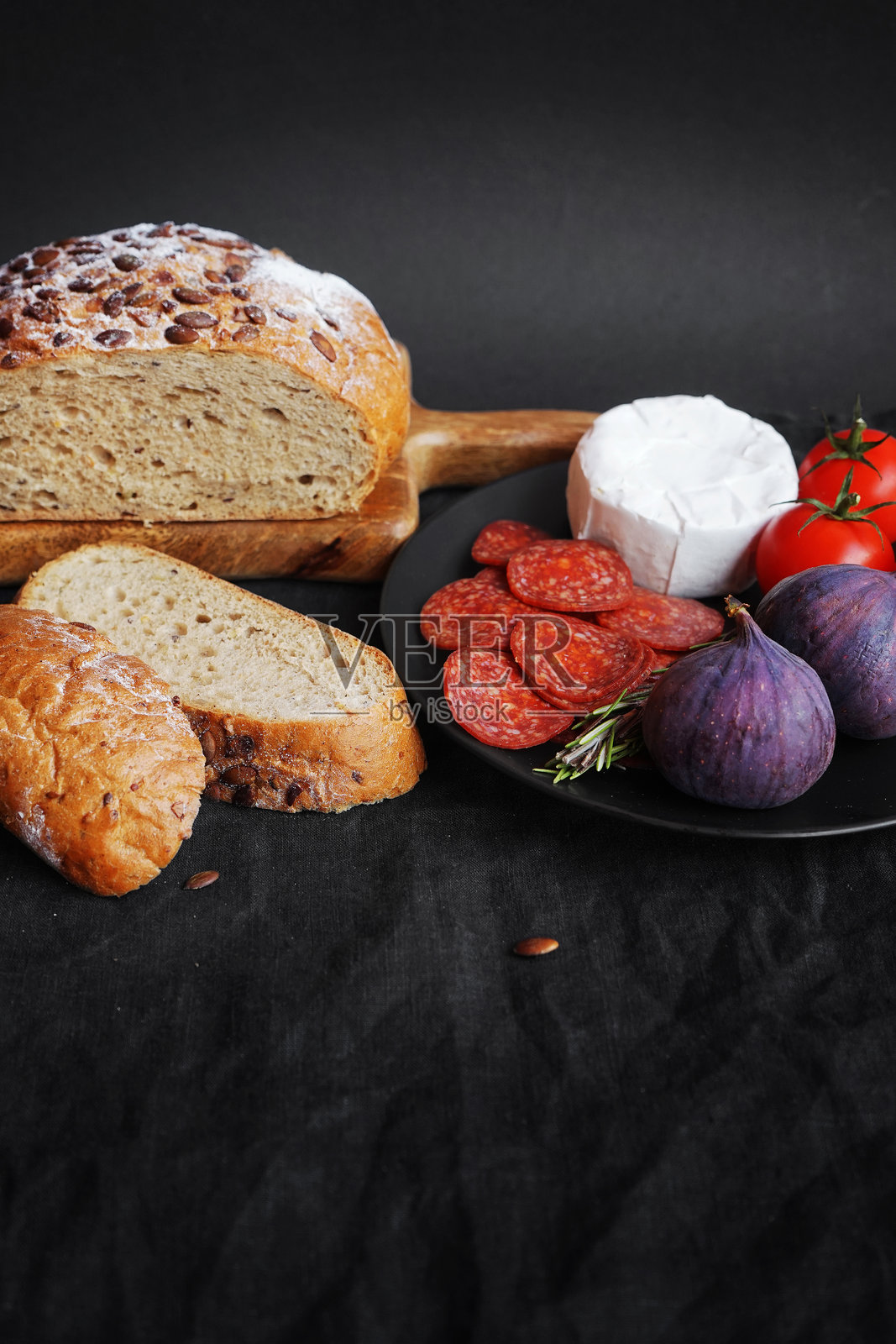 盘子里摆放着布里奶酪、佩罗尼香肠、西红柿、无花果和迷迭香枝,旁边是切片的全麦面包。照片摄影图片