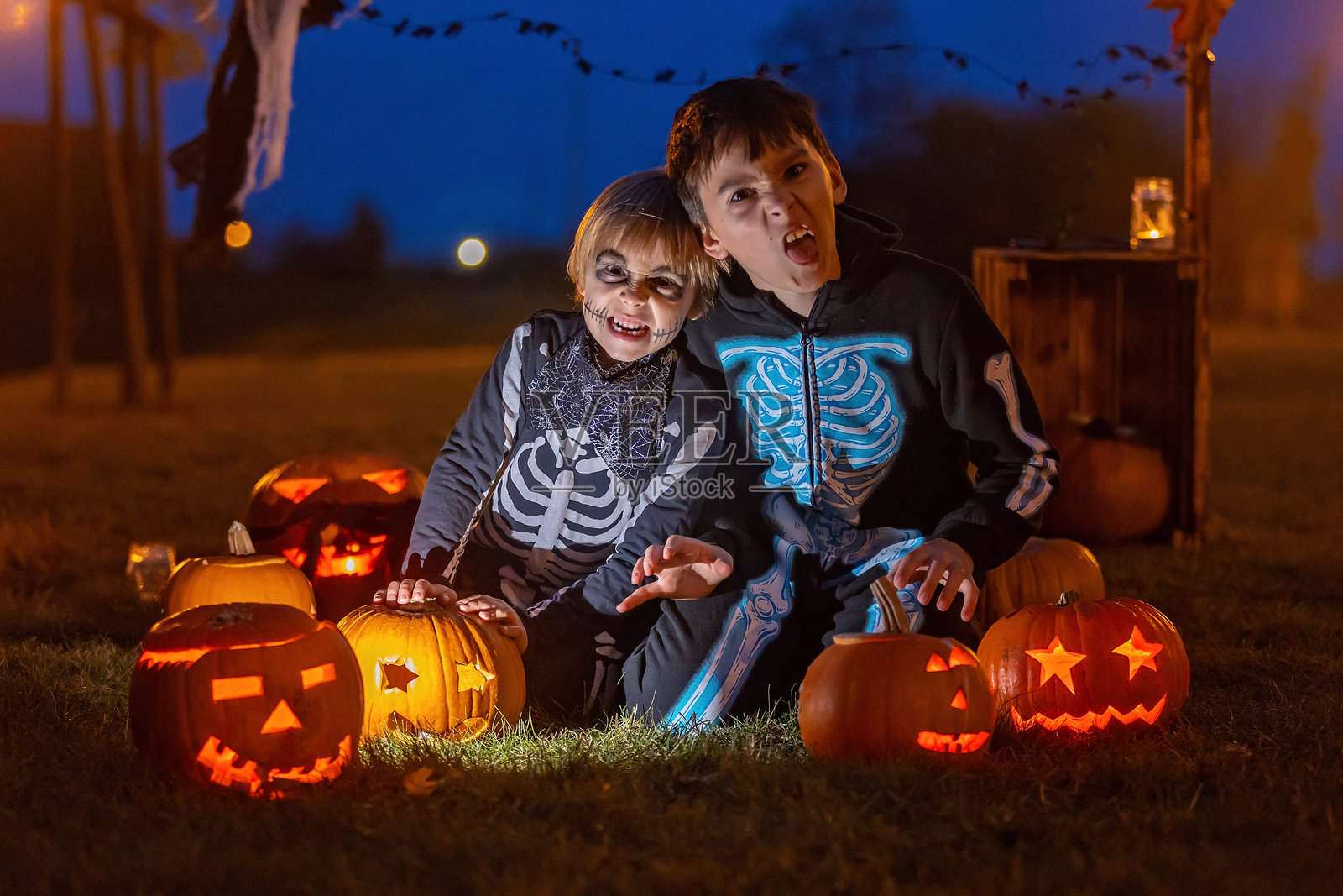 两个男孩在公园里穿着万圣节服装,旁边有雕刻的南瓜、蜡烛和装饰品。照片摄影图片