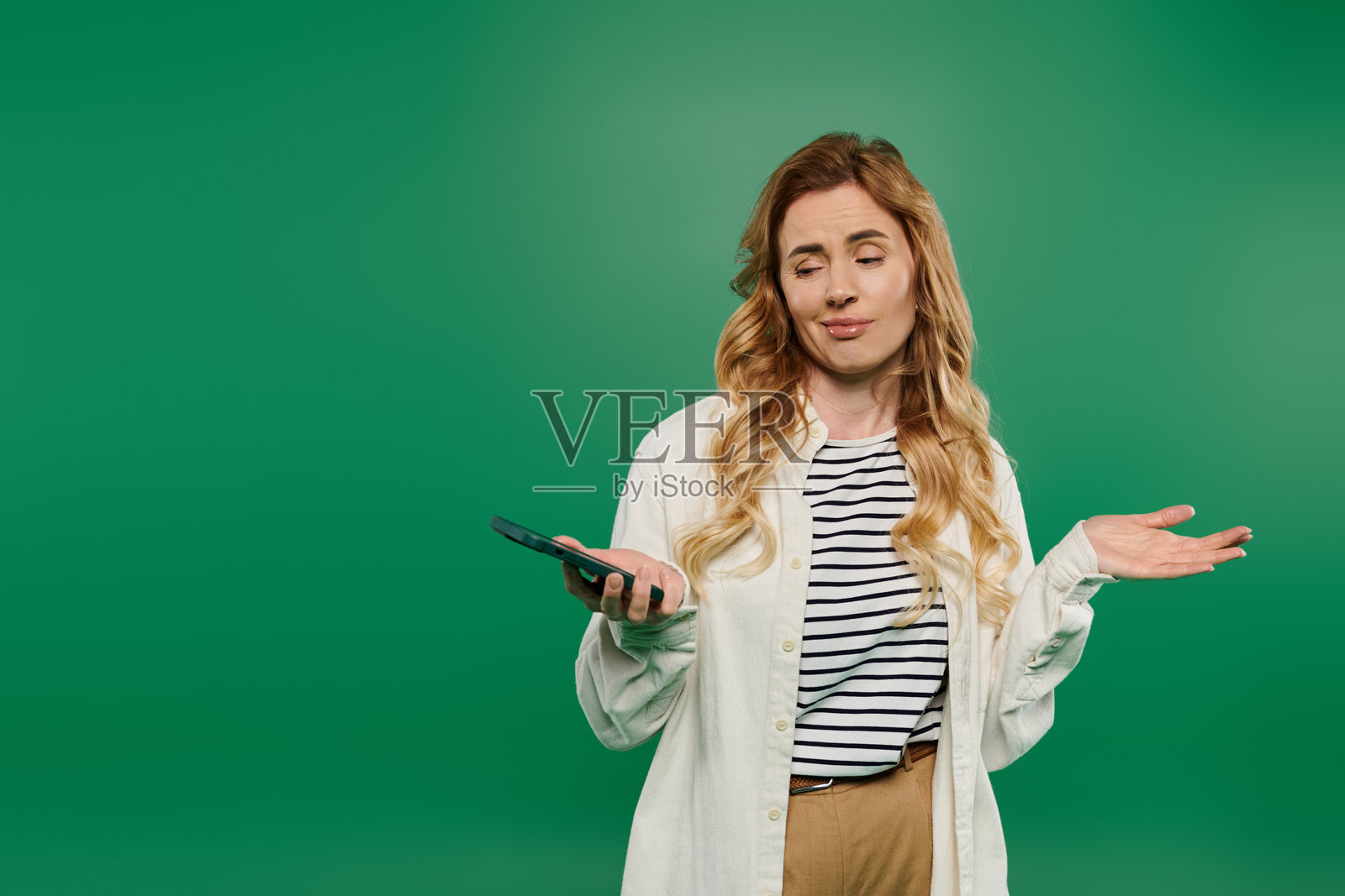 穿着休闲服的快乐女性在绿色背景前顽皮地摆姿势,手中拿着手机。照片摄影图片