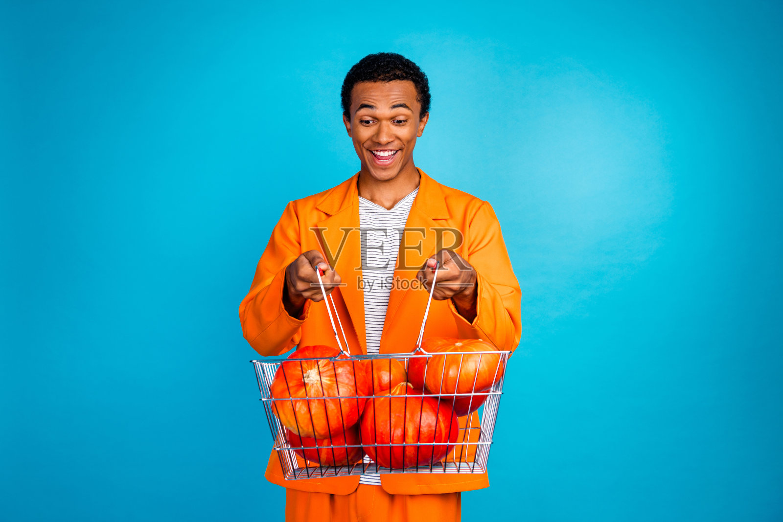 兴奋的男孩手持装满南瓜的超市购物篮,背景为蓝色,适合万圣节活动。照片摄影图片