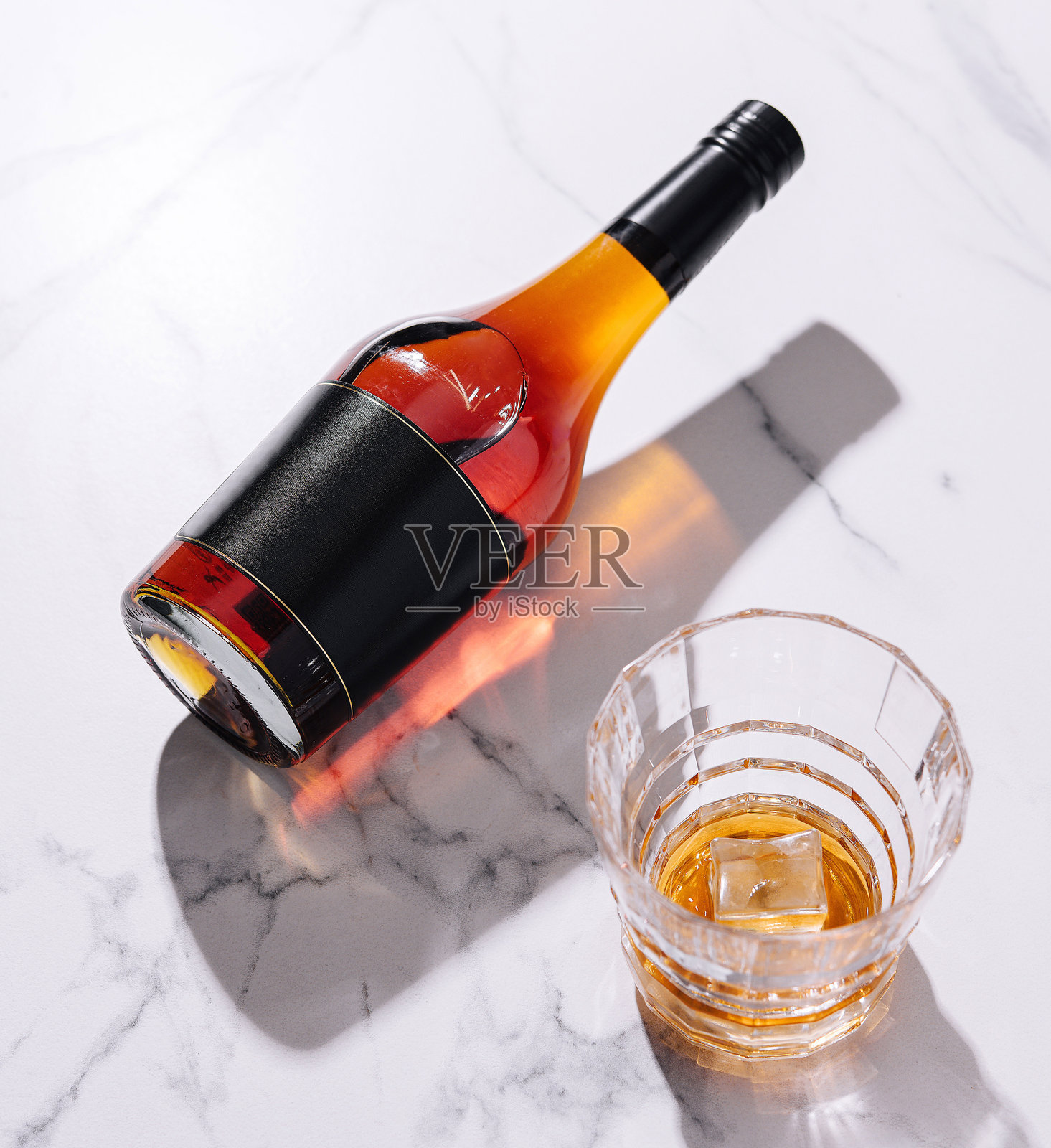 酒瓶在大理石上投下阴影,旁边是装有冰块的岩石杯照片摄影图片