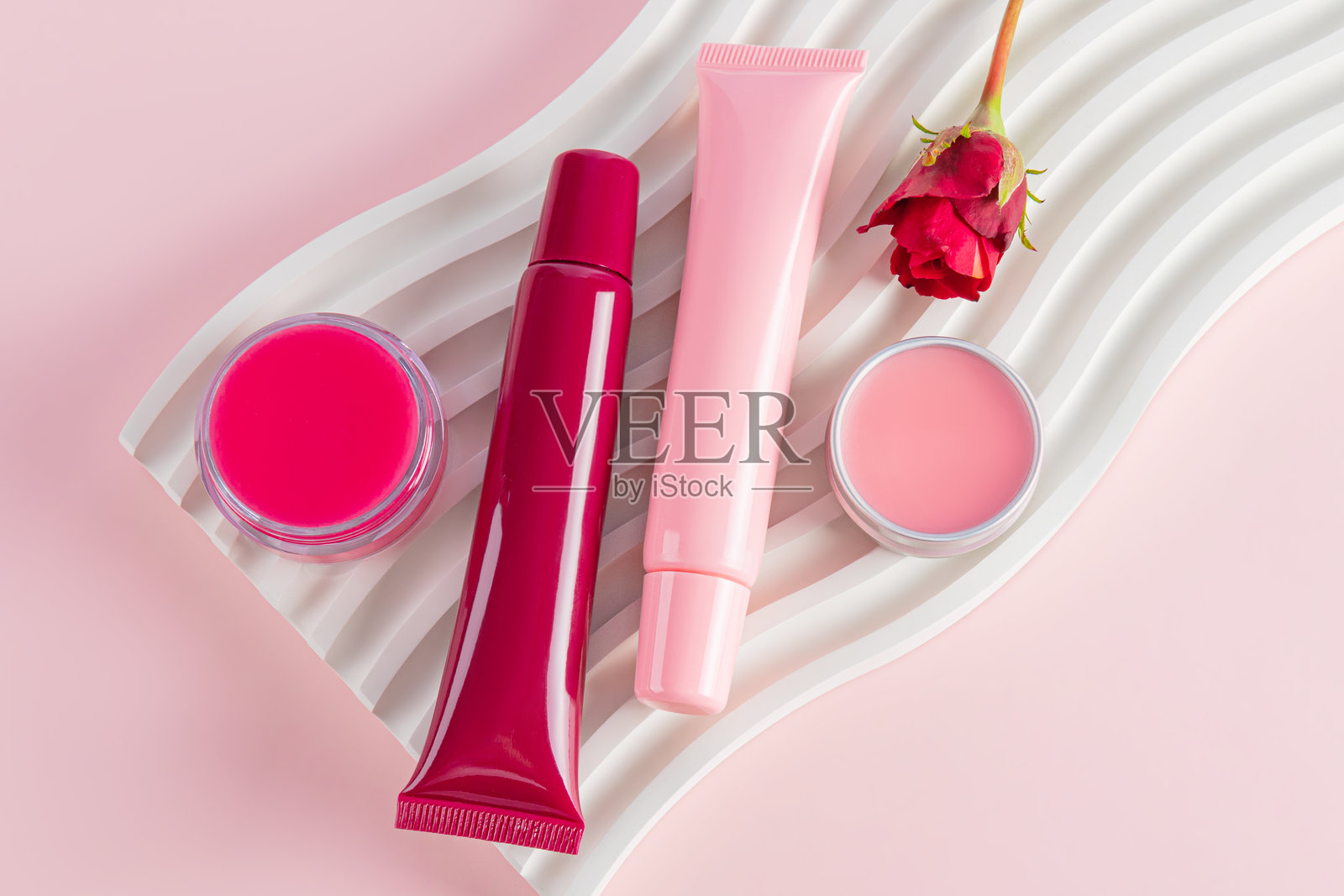 一组化妆品包装,包含多种类型的润唇膏和唇彩,置于波浪形的T台上。俯视图。粉色背景。自然护理。护理与美丽概念。照片摄影图片