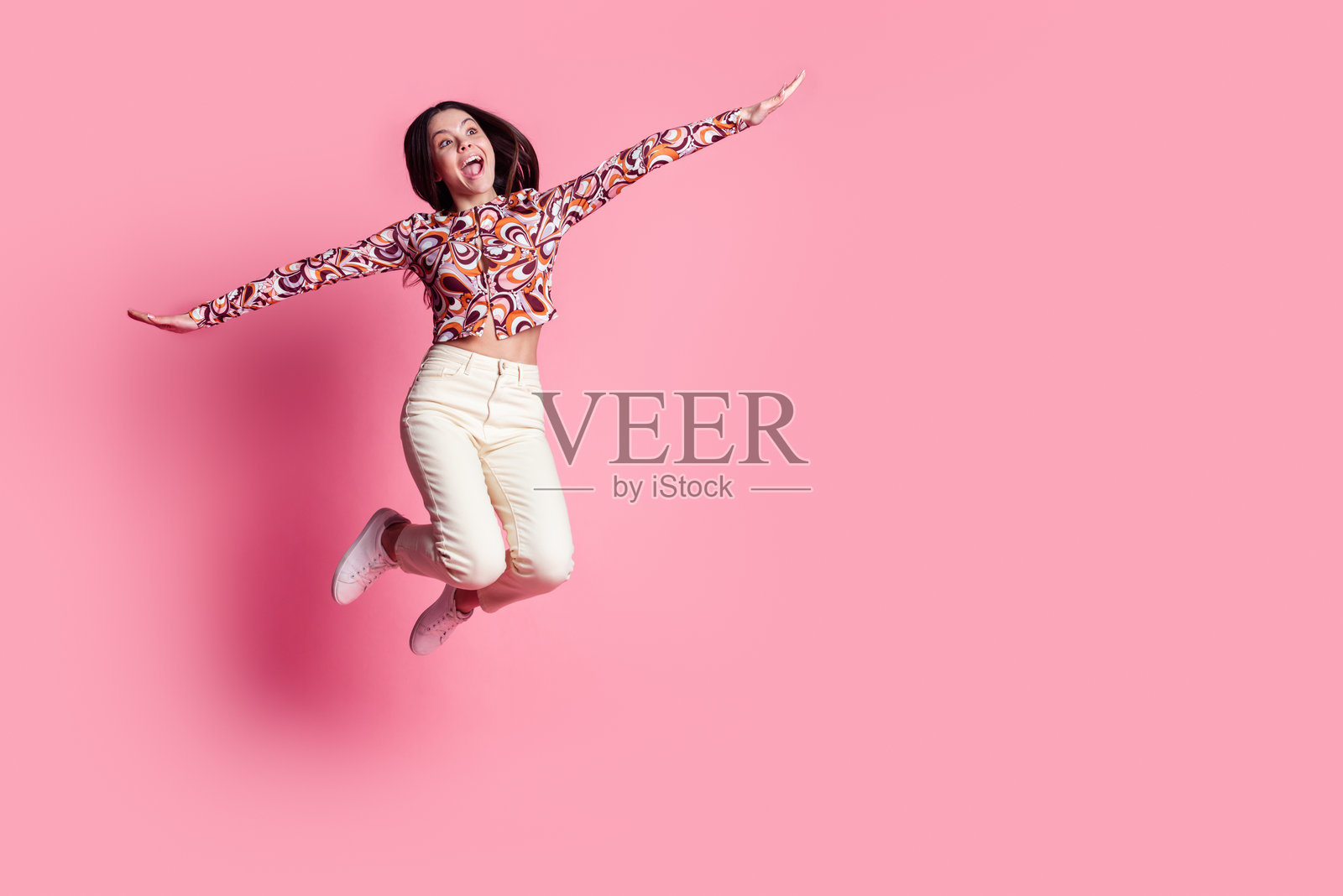 穿着时尚华丽服装的西班牙裔棕发少女在粉色背景上跳跃,像鸟一样飞翔。照片摄影图片