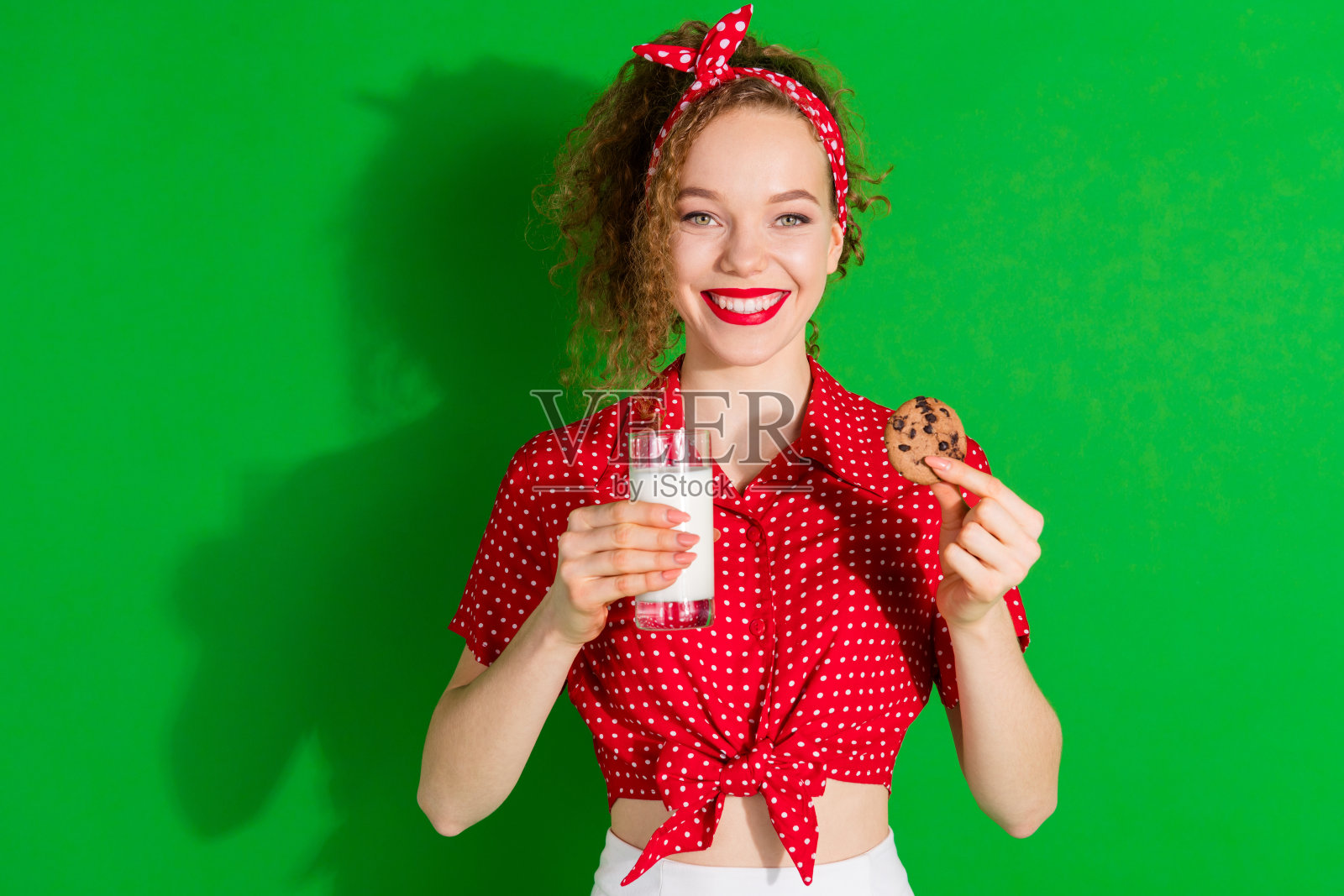 漂亮年轻女孩手持牛奶杯和饼干,穿着时尚的红色复古服装,背景为绿色。照片摄影图片