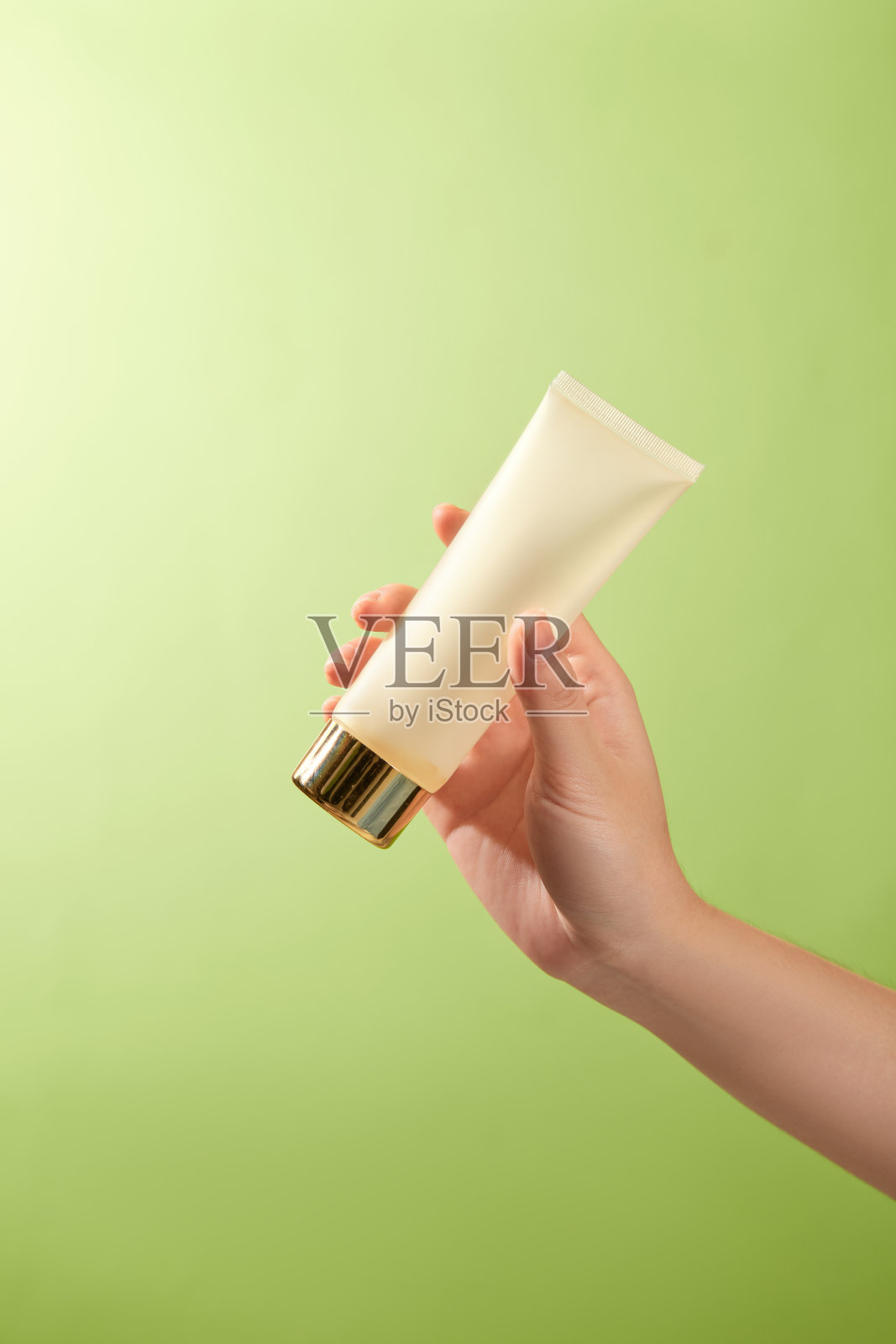 一只模特的手轻柔地握着一支无品牌的白色乳液管,配有金色盖子,展现出简约而优雅的设计。生动的绿色背景增强了产品自然清新的吸引力。照片摄影图片