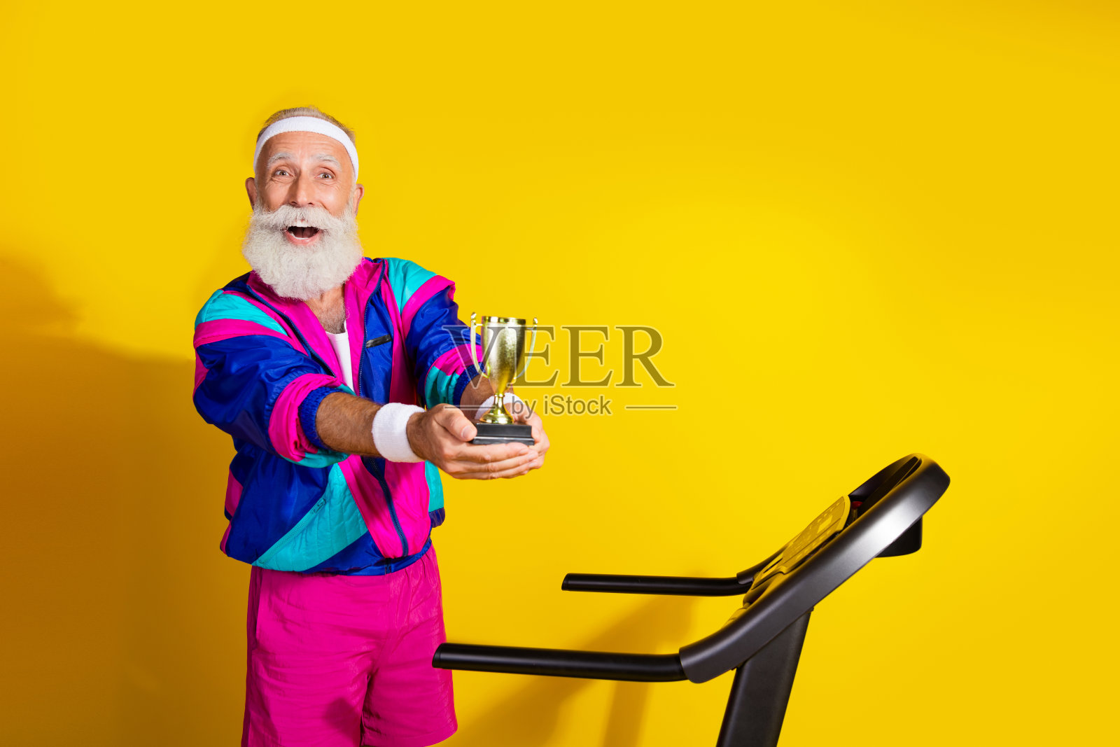 搞笑的老年运动员在黄色背景下穿着复古运动服,手持金色奖杯,站在跑步机上拍摄的肖像横幅。照片摄影图片