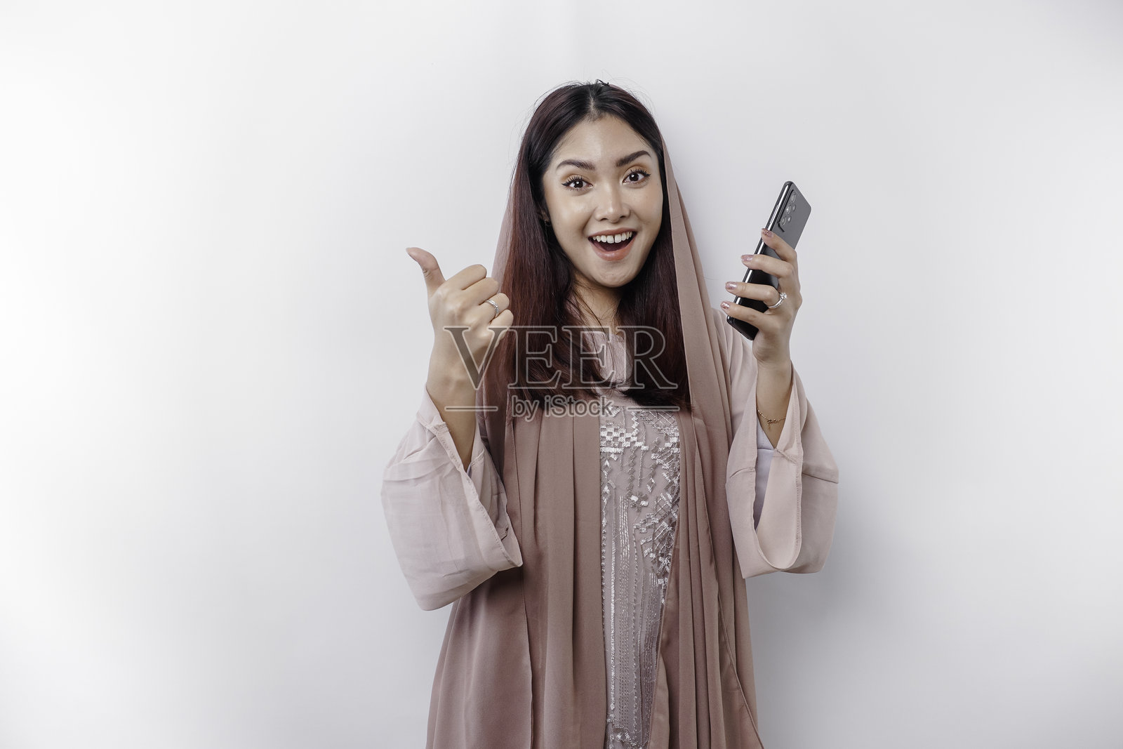 兴奋的亚洲穆斯林女性戴着头巾,竖起大拇指,同时手持智能手机。照片摄影图片