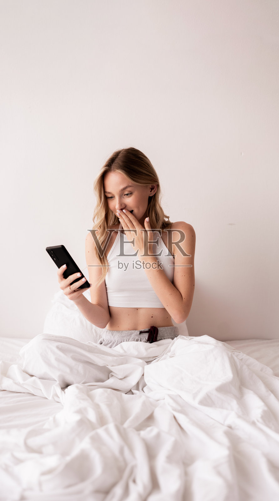 震惊的年轻白人女性坐在床上,因在智能手机上阅读新闻而感到震惊和惊讶。照片摄影图片