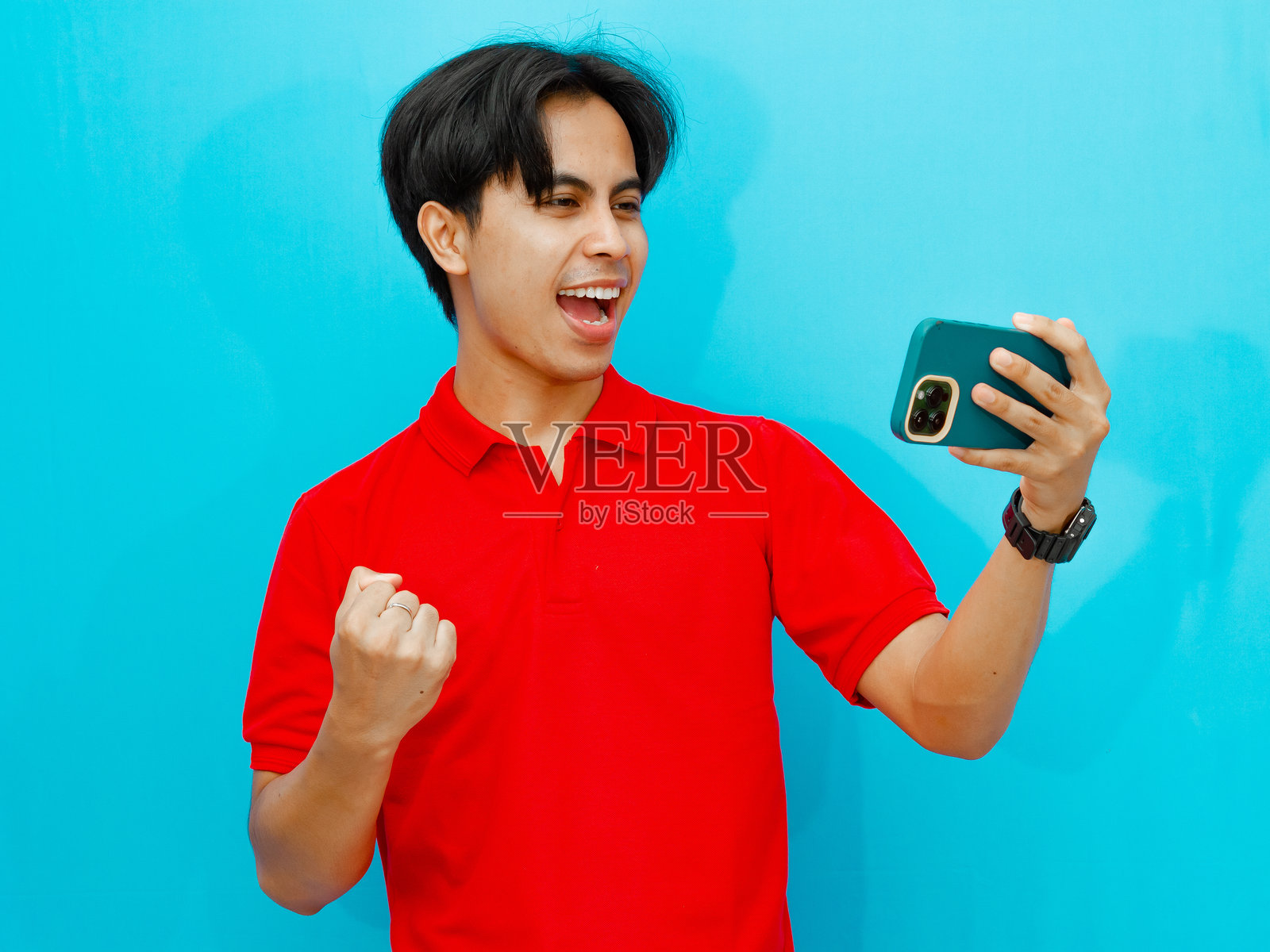 一位兴奋的亚洲男性玩家穿着红色 polo 衫,在玩手机游戏时庆祝胜利。背景为蓝色,展现出热情和成功。照片摄影图片