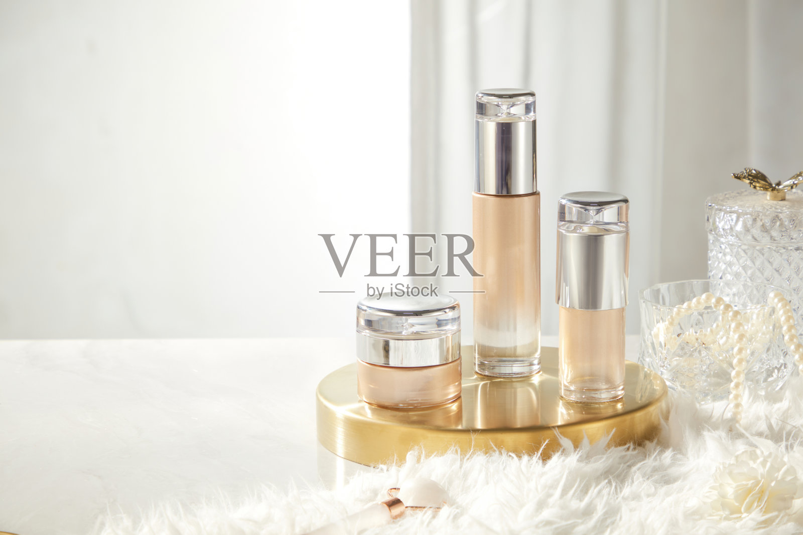 一张用于推广美容产品的图片,展示了一个罐子和两个没有标签的瓶子,放置在一个铜托盘上,周围有一块毛皮地毯,玻璃瓶上缠绕着珍珠串。照片摄影图片