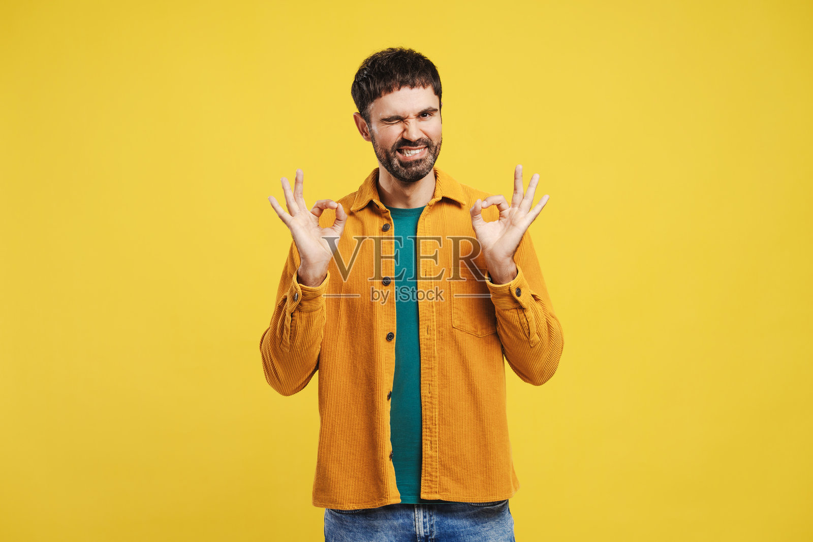迷人的拉丁男子眨眼,做出“好”的手势,穿着时尚的黄色衬衫,正看着镜头。照片摄影图片