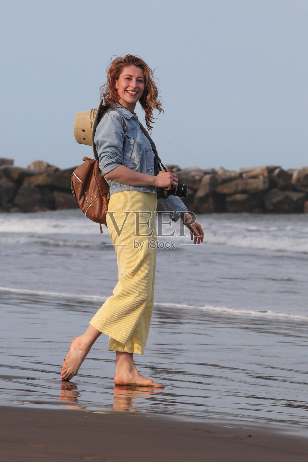 在日落前,背着背包和相机的快乐女性在海滩上。照片摄影图片