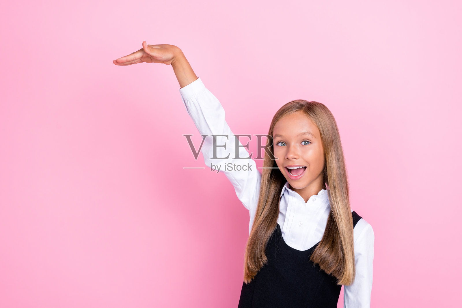 穿着时尚衣服的快乐漂亮女孩站在空旷的地方,展示她想要多高,背景为粉色。照片摄影图片