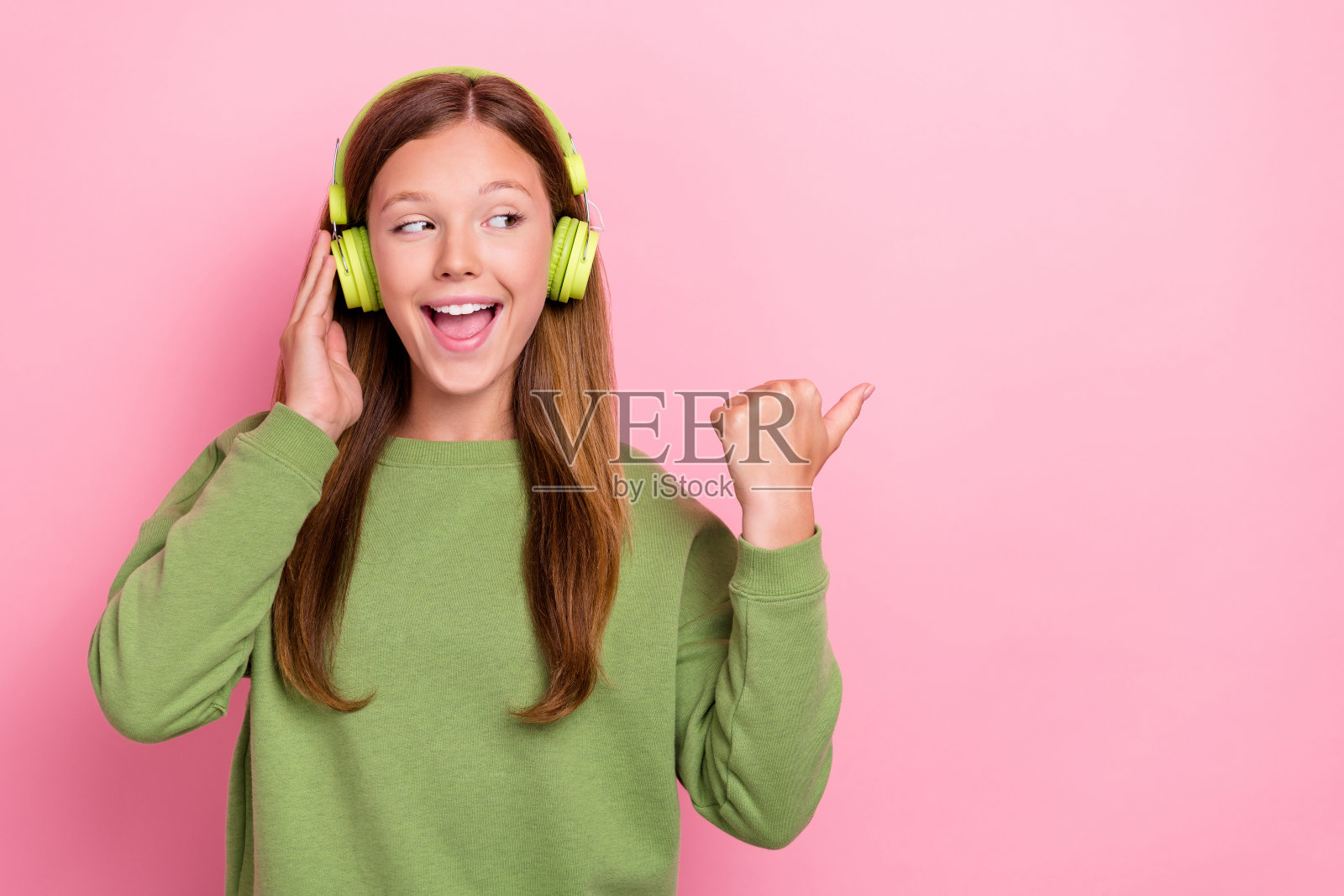 穿着绿色卫衣、戴着耳机的可爱兴奋的女学生,拇指指向空白处,背景为粉色。照片摄影图片