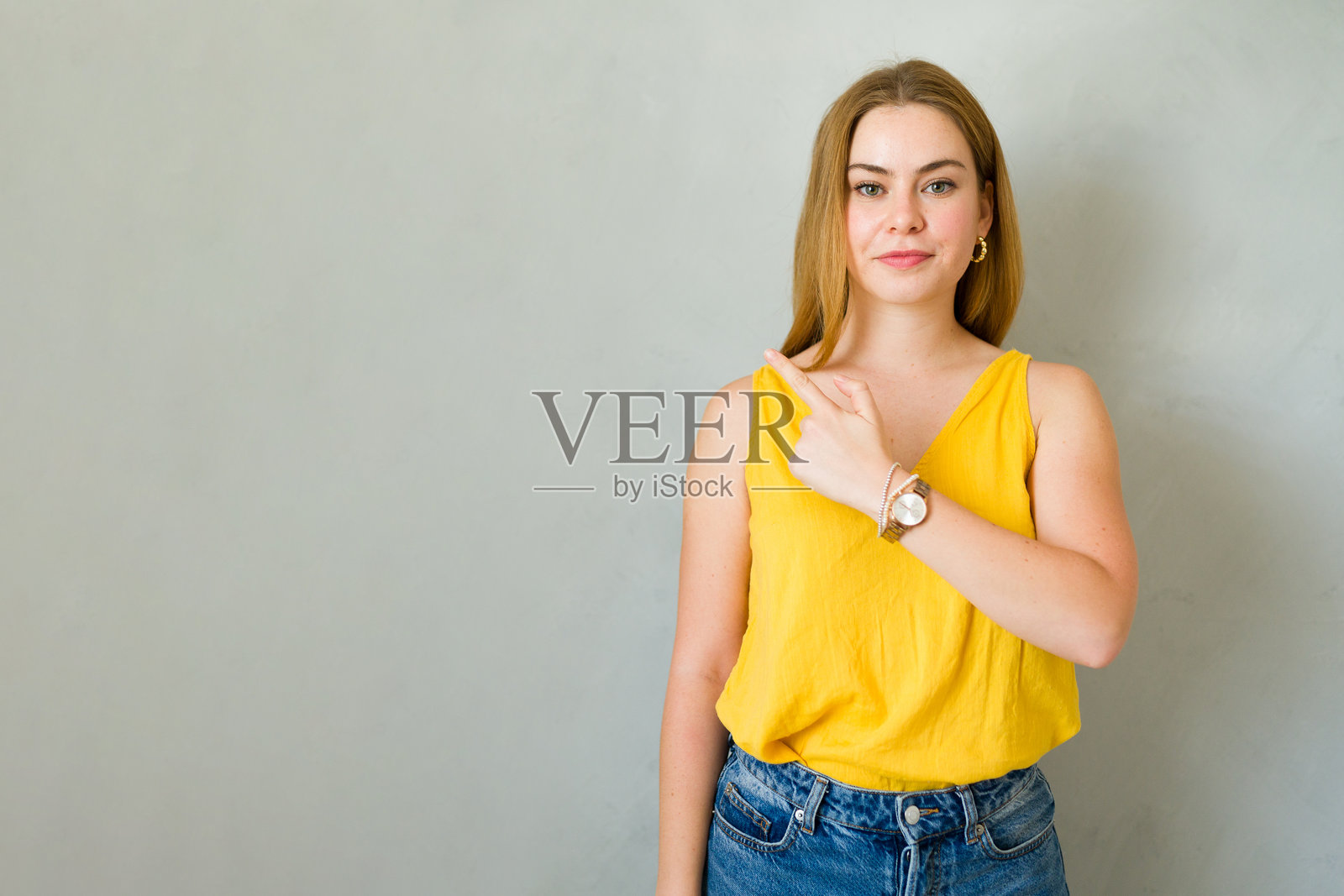 可爱的年轻女性用食指指向右侧,背景为灰色。照片摄影图片