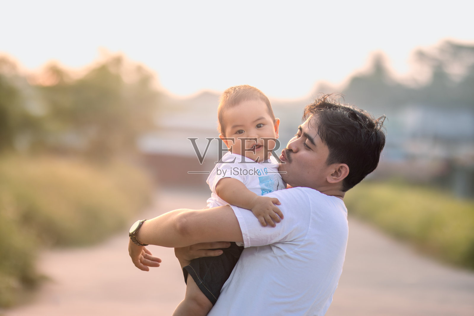 父亲抱着婴儿男孩,想要亲吻他。婴儿男孩对着镜头露出天真的微笑。父亲和婴儿男孩都穿着可爱的白色T恤。照片摄影图片