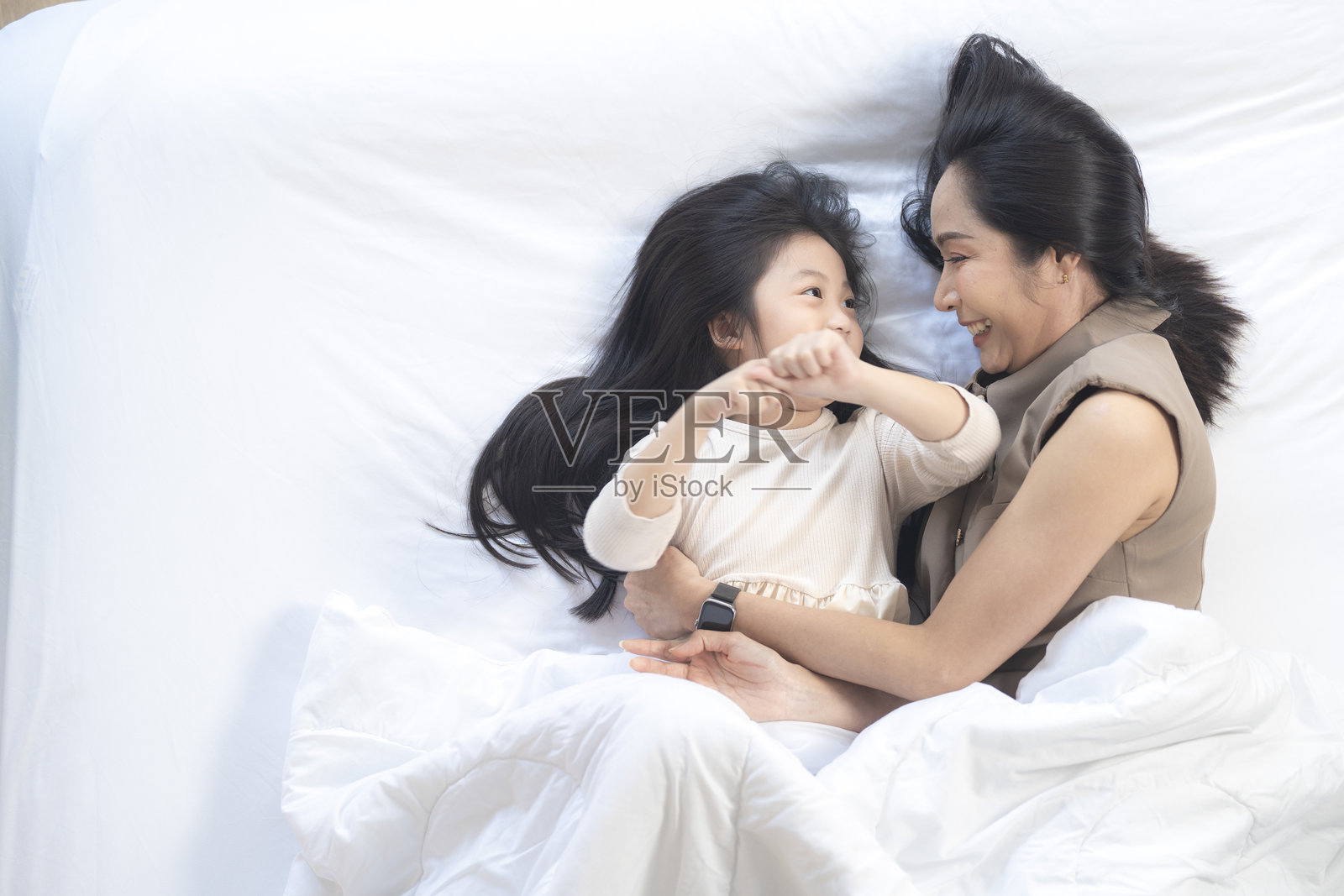 一位快乐的母亲和女儿躺在床上一起欢笑。这个愉快的时刻展现了她们之间的深厚纽带和在舒适家居环境中的幸福,周围环绕着自然装饰。照片摄影图片