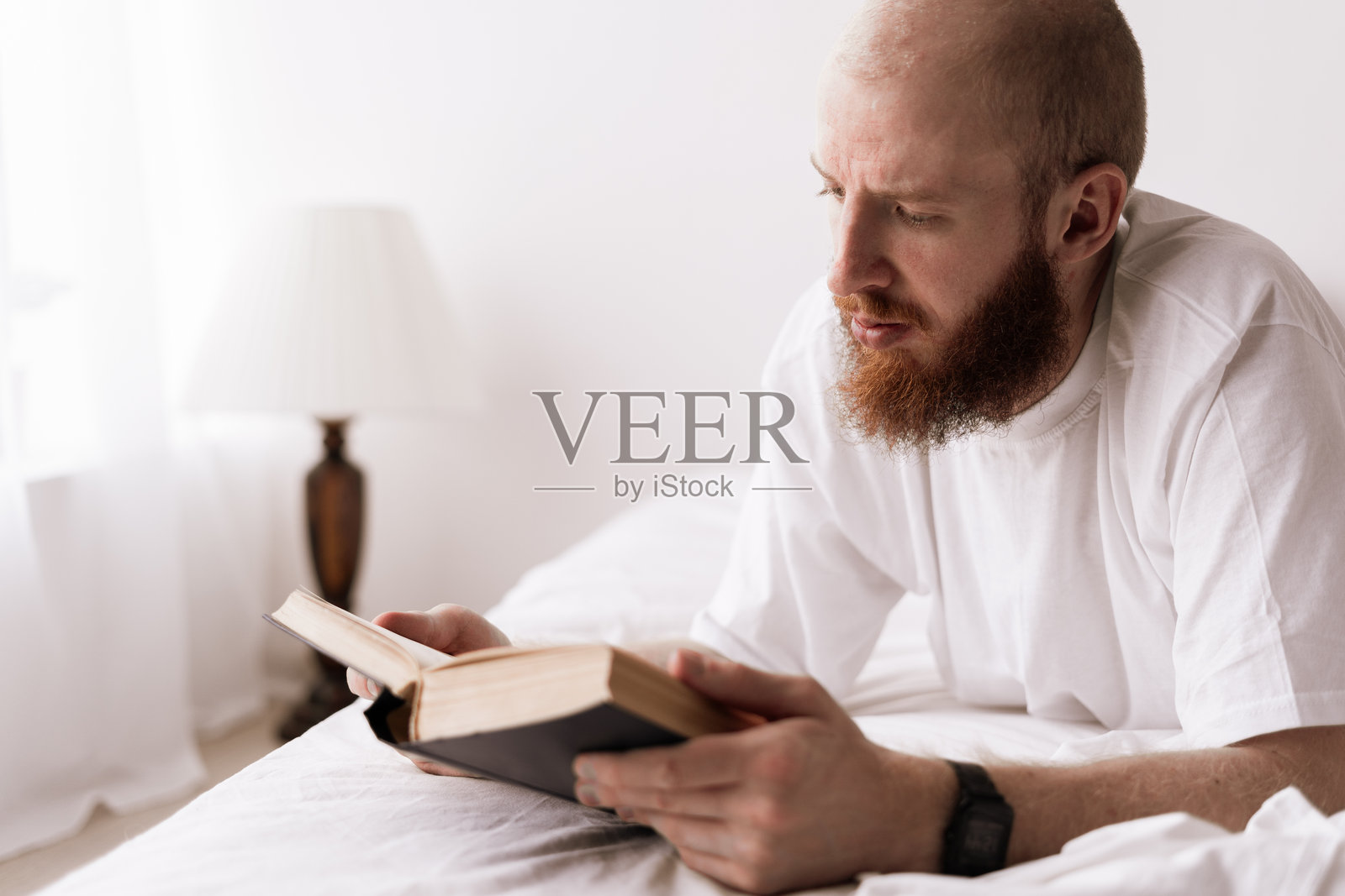 留着胡子的男人在卧室的舒适床上阅读一本书。白人男性在休息日享受畅销书的乐趣。照片摄影图片