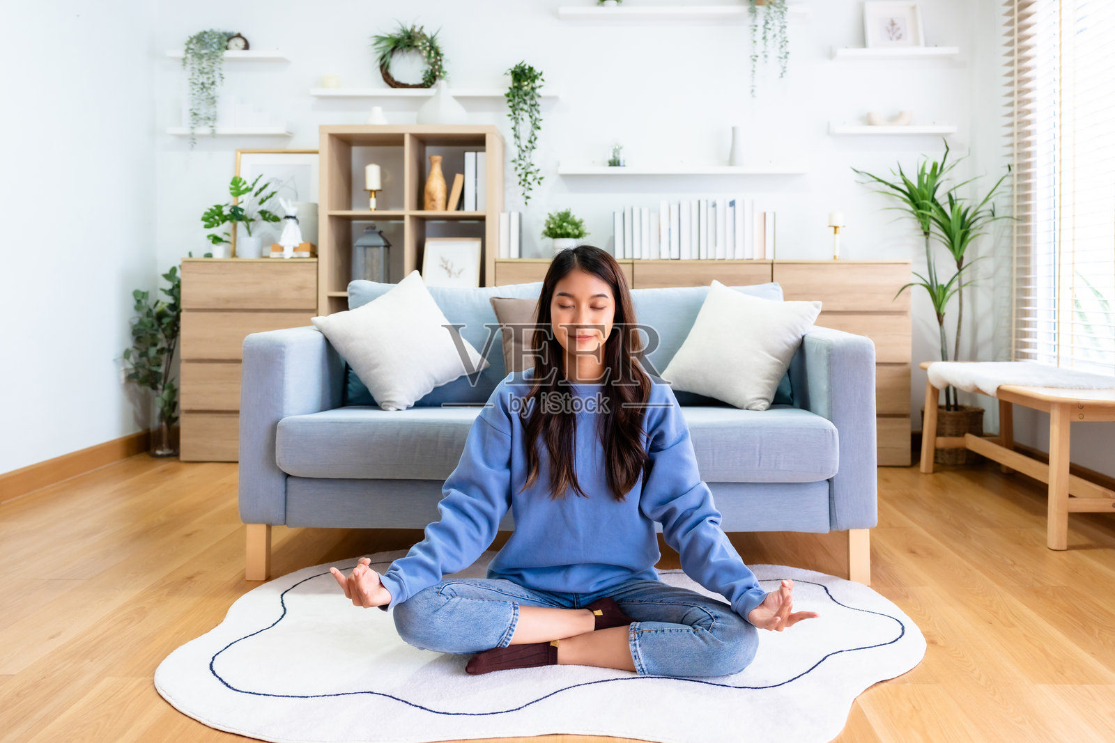 快乐的年轻亚洲女性在家中练习瑜伽和冥想,坐在客厅的地板上,双腿交叉,闭眼放松。正念冥想与身心健康的概念。照片摄影图片