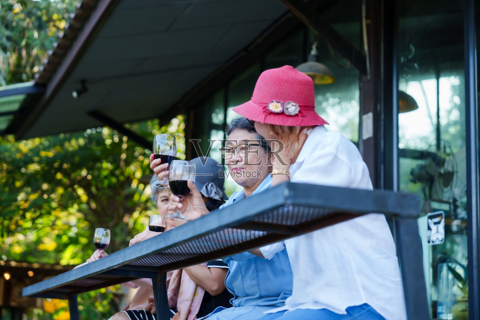 一群亚洲老年女性坐在餐厅的户外座位区,享受红酒。她们穿着休闲,气氛轻松,面带愉快的笑容,背景中有绿意,展现了社交的氛围。照片摄影图片