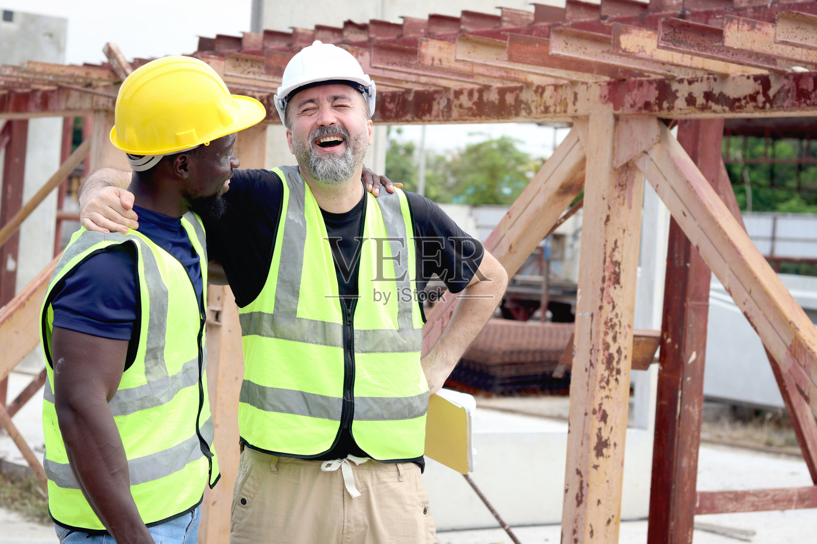快乐和谐的职场人们,微笑的白人工头和非洲工程师在建筑工地上亲密相拥。种族肤色的差异中充满爱与尊重,体现多样性。照片摄影图片