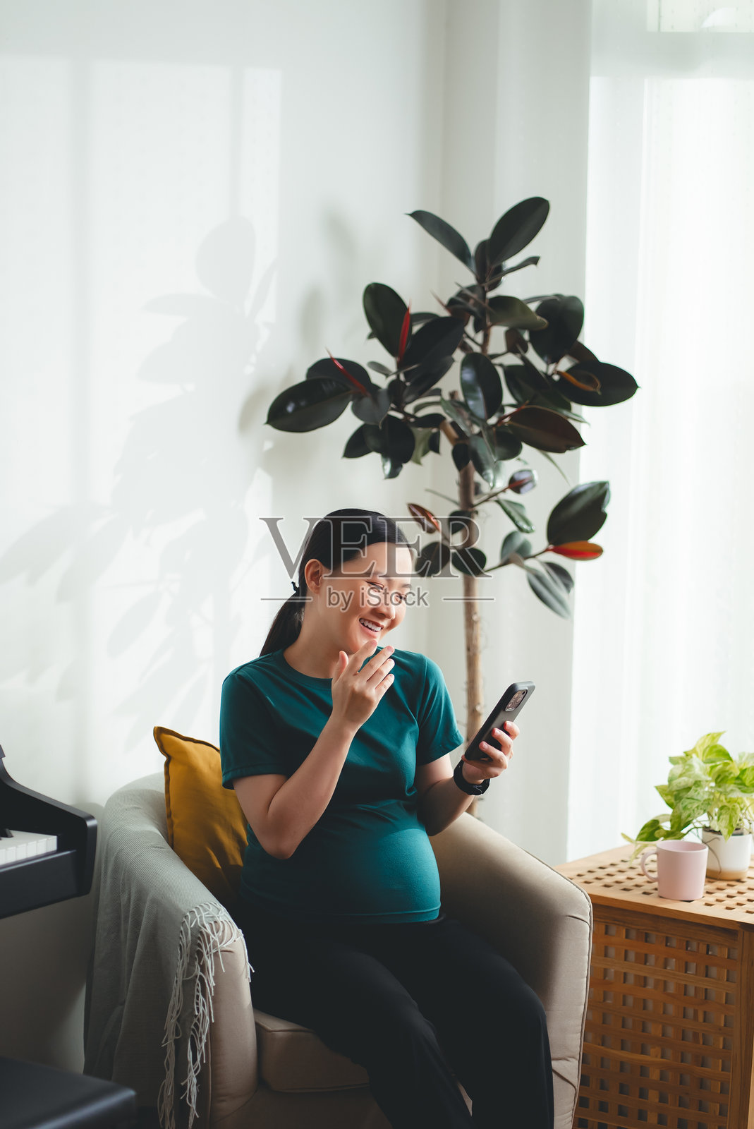 亚洲孕妇坐在扶手椅上开心地微笑,正在家中使用智能手机。照片摄影图片