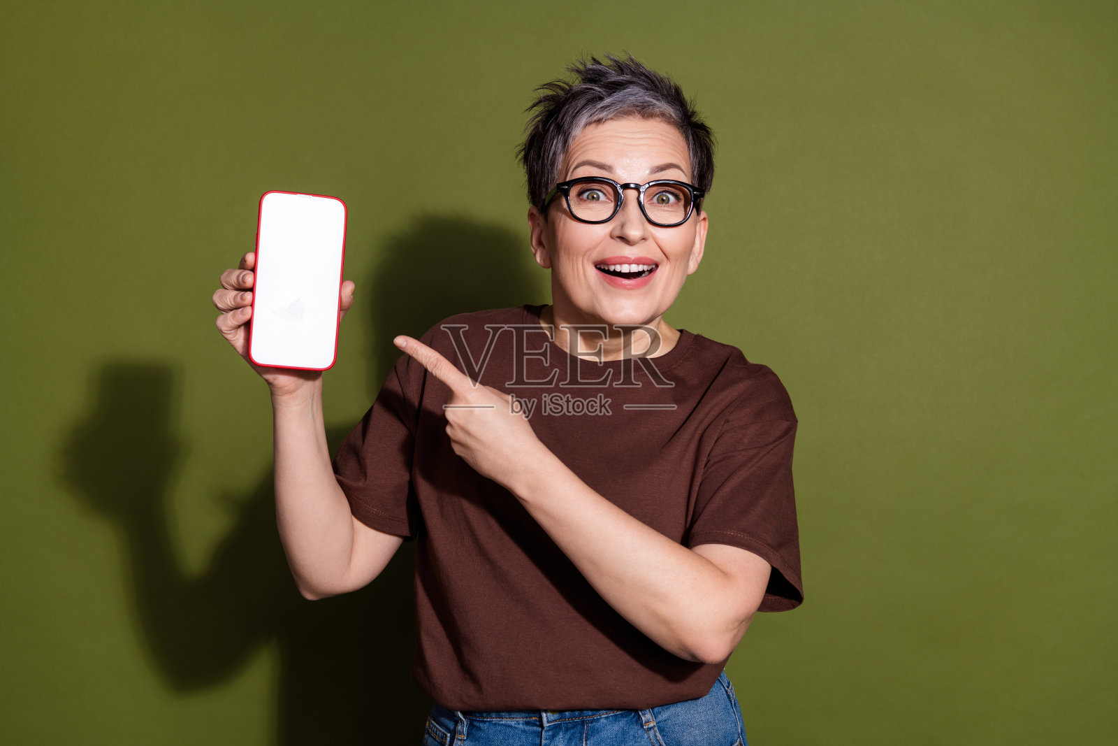 漂亮的老年女士指着空白的智能手机屏幕,穿着棕色T恤,背景为卡其色。照片摄影图片