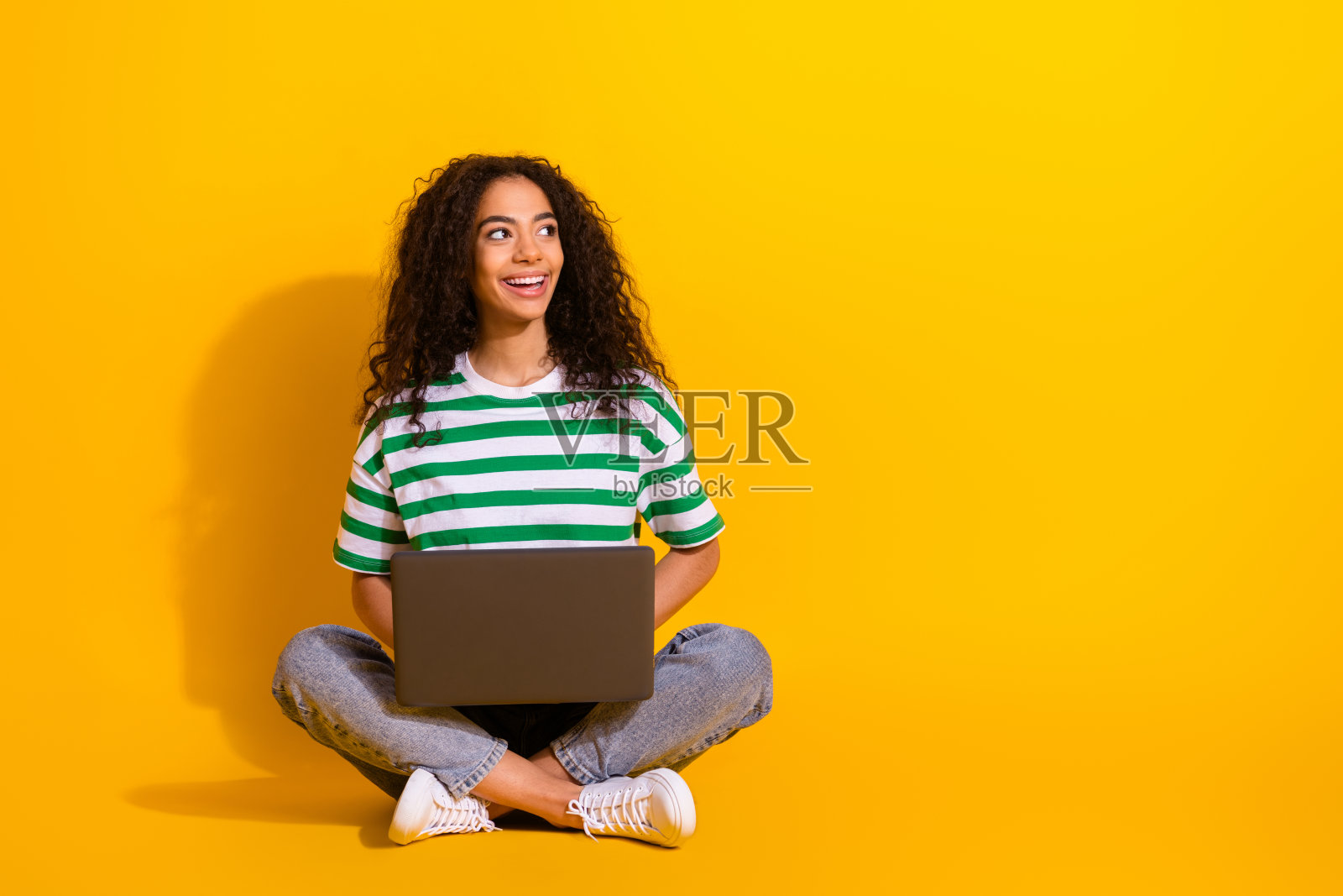 全身肖像,年轻女孩穿着条纹T恤,坐在笔记本电脑前,面带微笑,背景为黄色,显得空旷。照片摄影图片