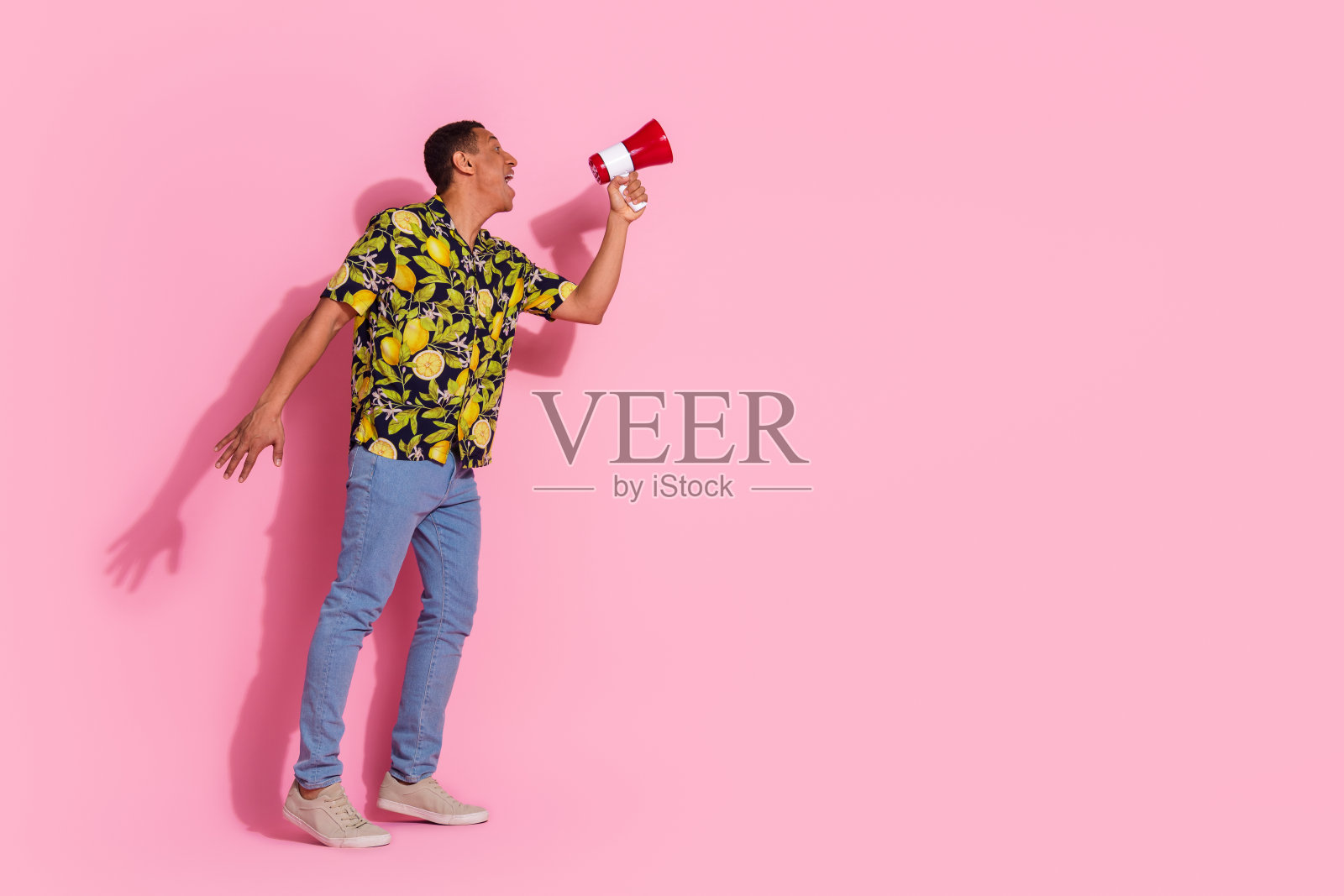 全身照,年轻帅气的男孩用扩音器大喊,穿着时尚的热带印花服装,背景为粉色。照片摄影图片