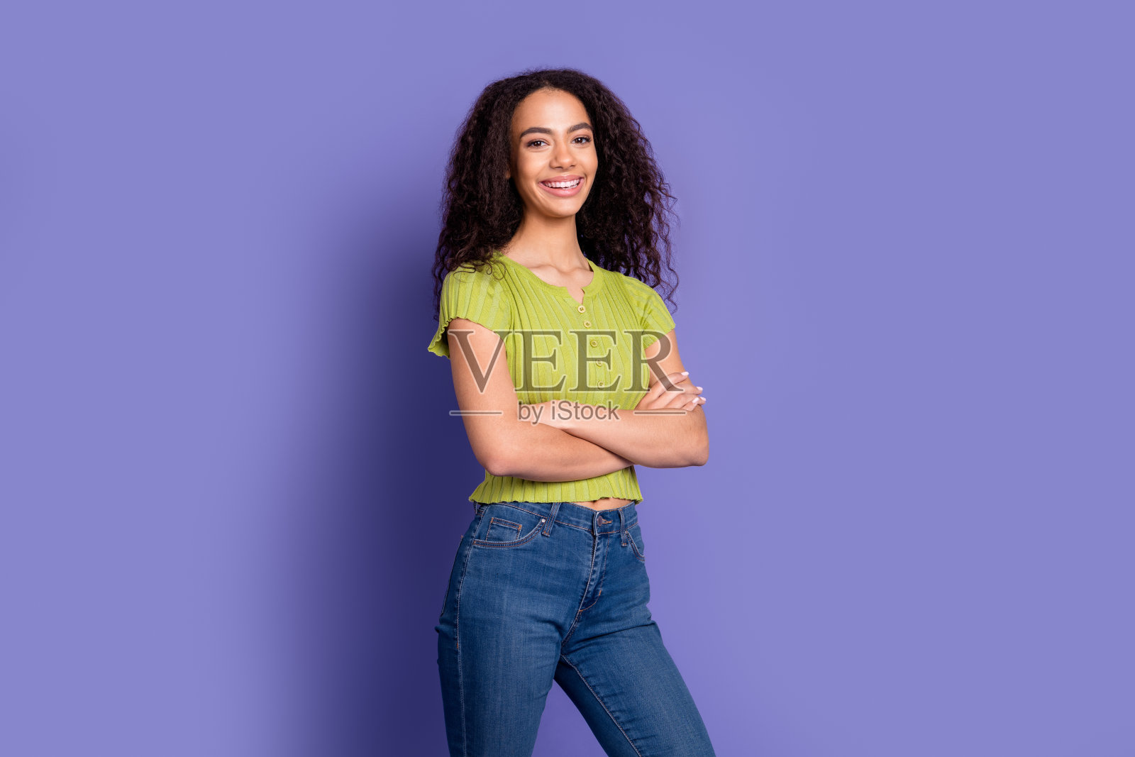 可爱的少女自信地交叉双手,穿着时尚的绿色服装,背景为紫色。照片摄影图片