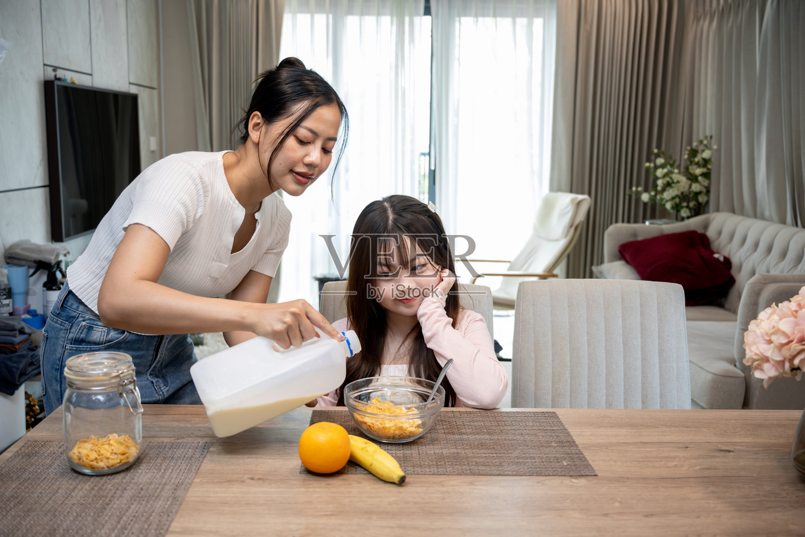 一个亚洲女孩坐在餐桌旁,她对早餐的麦片感到兴奋。她的母亲正在为女儿从一加仑的牛奶中倒牛奶。照片摄影图片