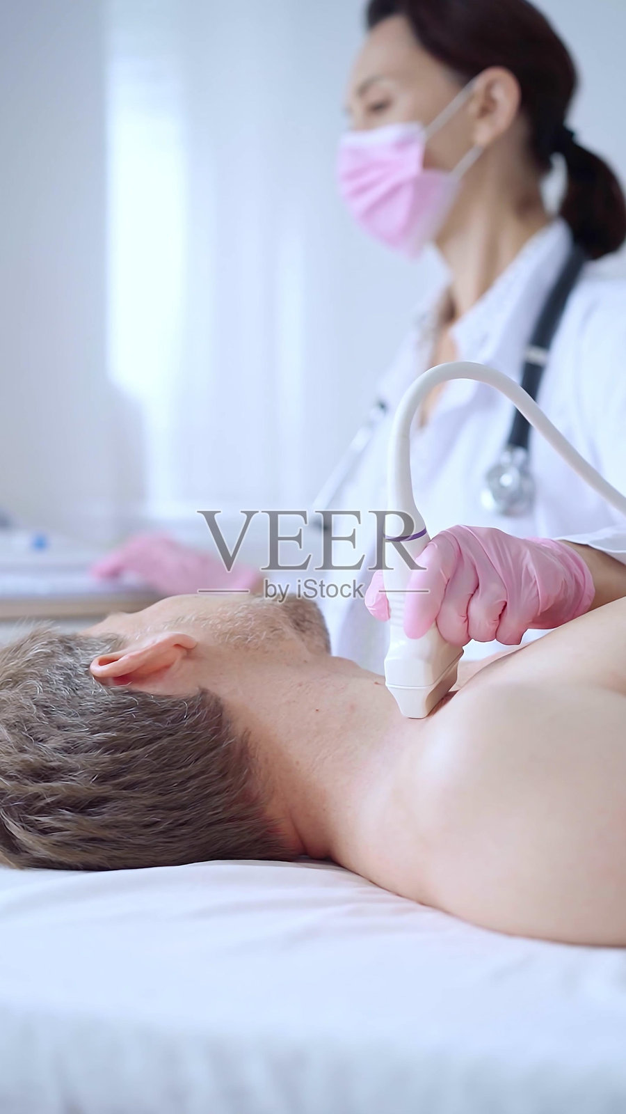 戴着粉色医用手套和口罩的女医生为男病人进行颈部超声检查,甲状腺检查,医学概念照片摄影图片