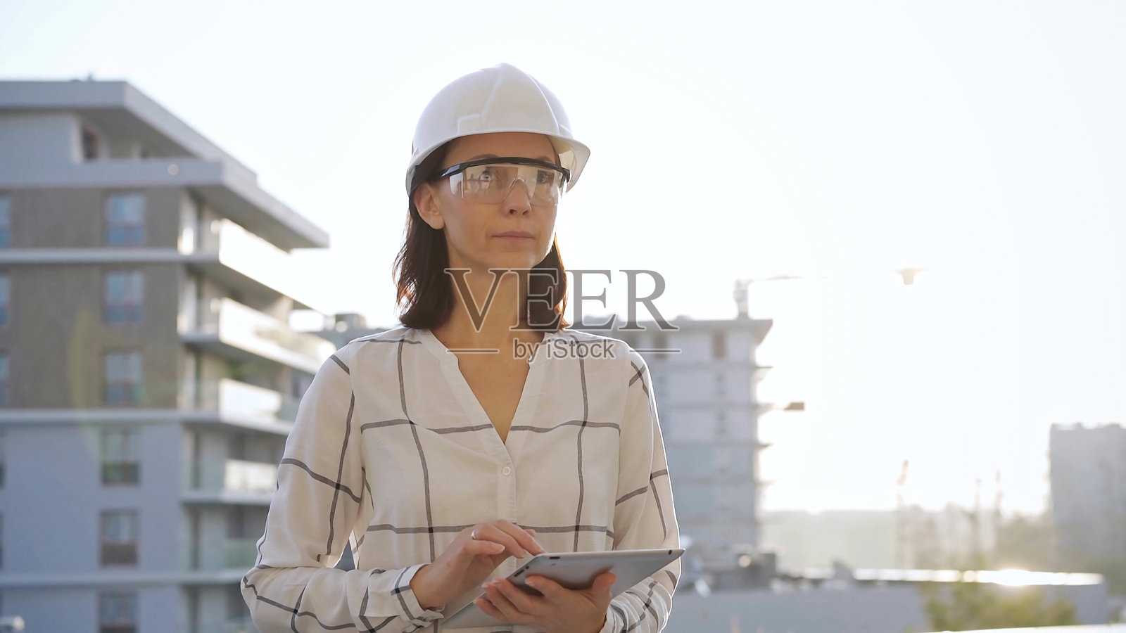 穿着白色格子衬衫和安全帽的女性建筑工程师在日落时分检查建筑工地,手持平板电脑,正面视角。建筑和工程概念照片摄影图片