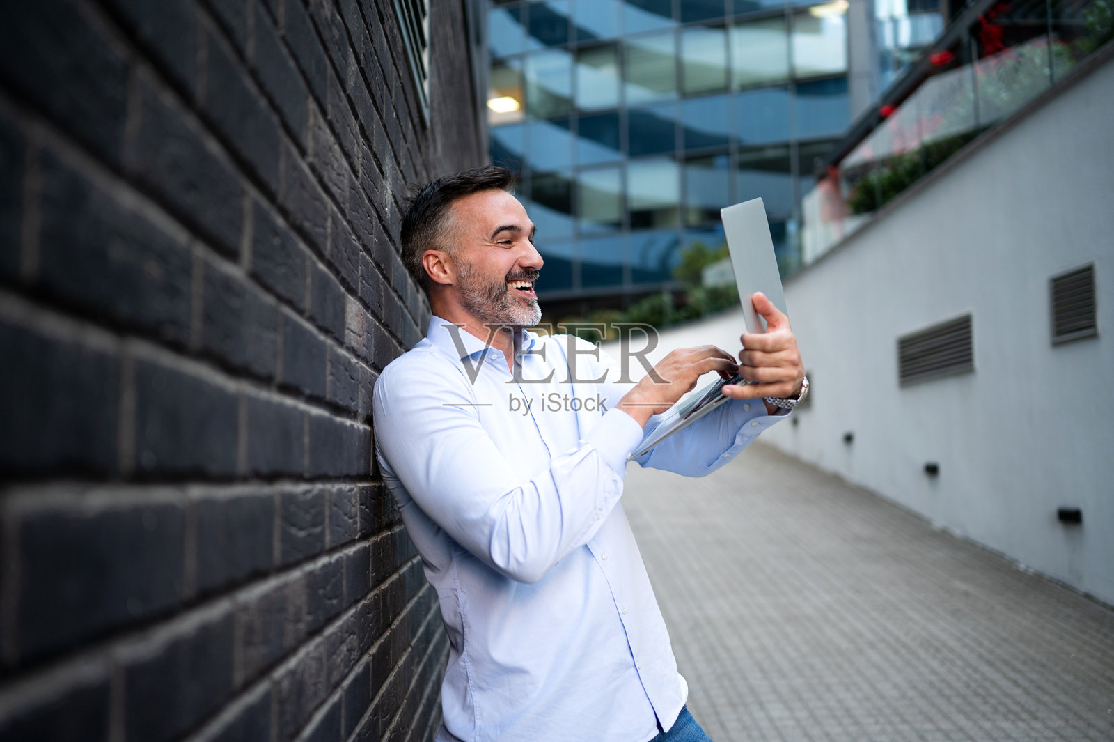 中年商人靠在墙上,在空荡荡的走廊里使用笔记本电脑照片摄影图片