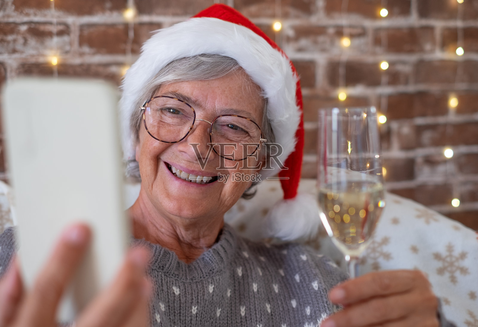 微笑的白人老年女性穿着冬季毛衣,坐在家中,手持香槟杯,一边用手机视频聊天与家人或朋友庆祝圣诞节和新年。照片摄影图片