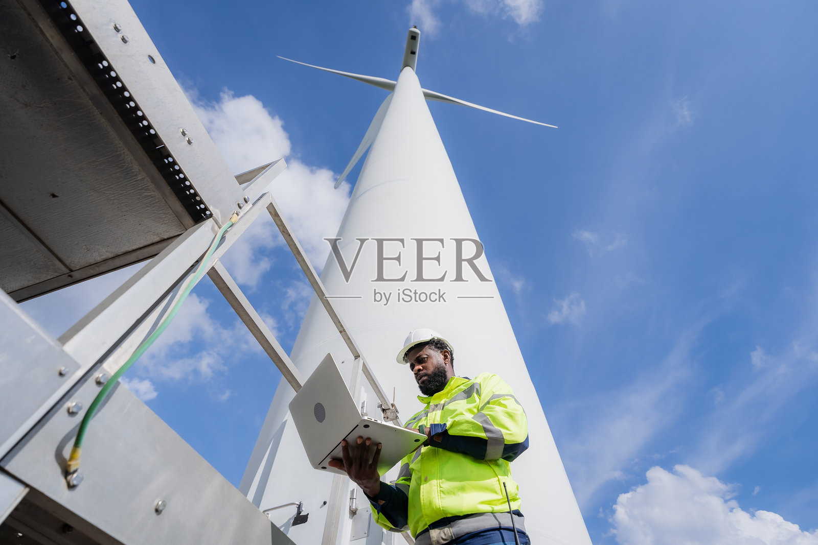 一名穿着黄色夹克的男子正在检查风力涡轮机。他正在使用笔记本电脑查看涡轮机的状态。电气,工程师,环境,能源,清洁,可持续,风力涡轮机,自然照片摄影图片