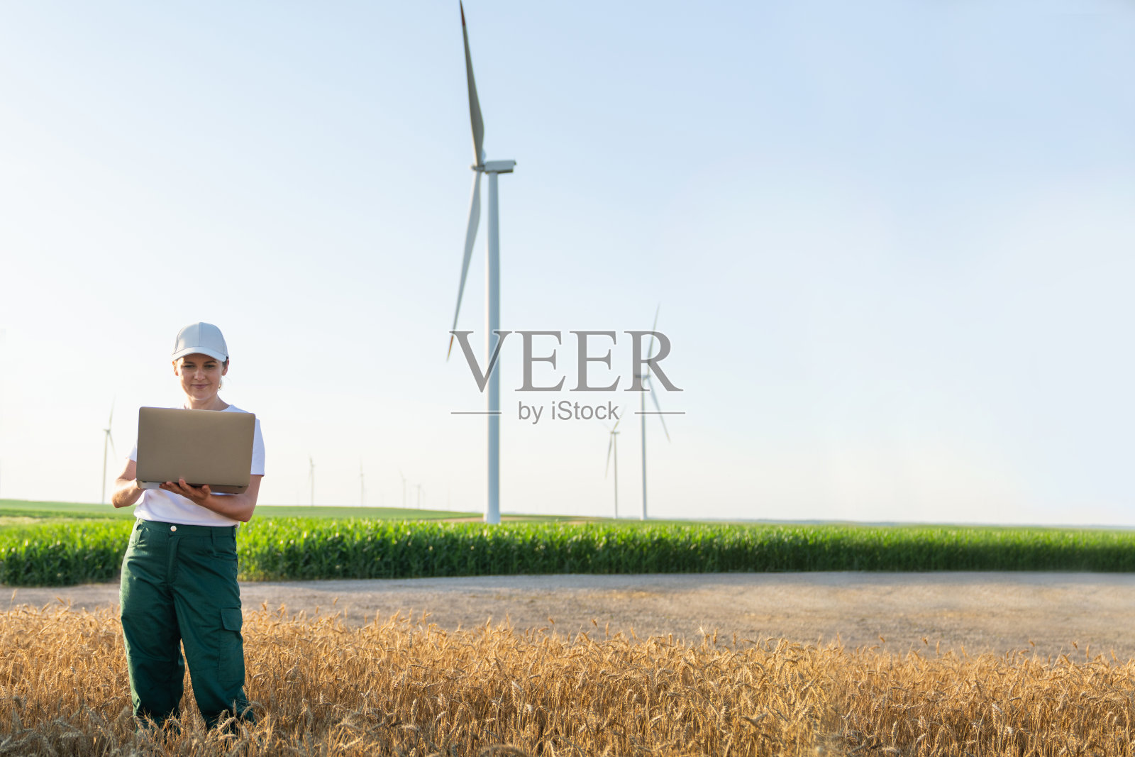 穿着白色帽子和T恤的女性农民手持笔记本电脑,站在日落时分的农田中,背景有风力发电机。留白空间。照片摄影图片