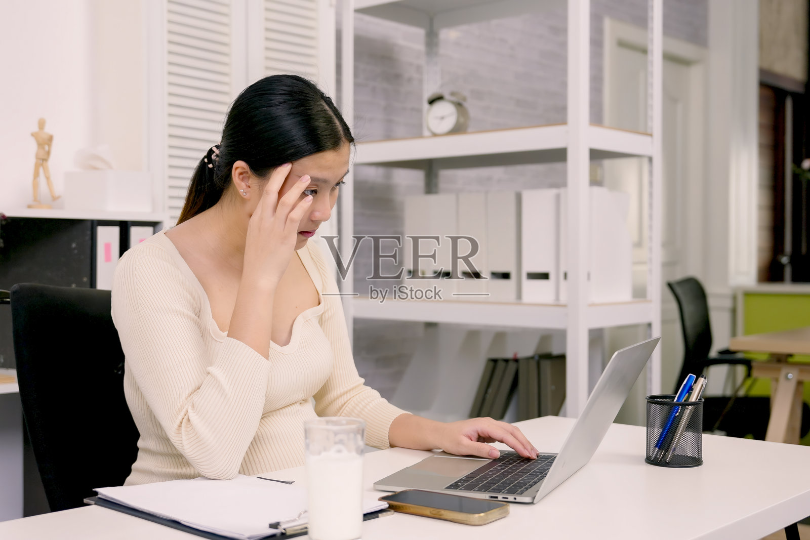 在办公室椅子上的怀孕亚洲妇女,手放在额头上,在专业的家庭办公室环境中工作时,表现出工作与生活平衡的挑战照片摄影图片