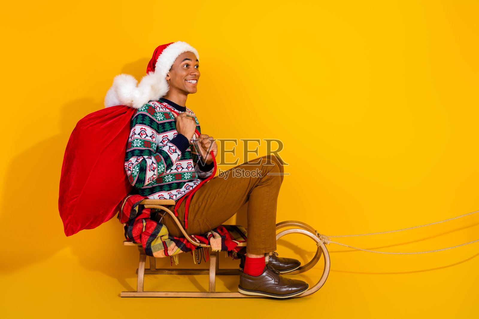 年轻男子全身照,手持礼物袋,坐在空的雪橇上,背景为黄色,充满圣诞节的节日气氛。照片摄影图片