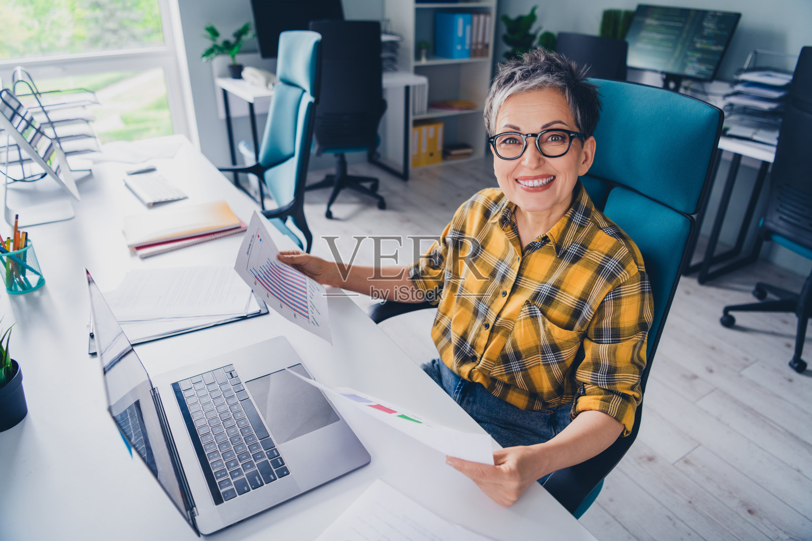 可爱的退休女士在舒适的初创办公室里穿着格子黄色衬衫,处理文件和图表。照片摄影图片