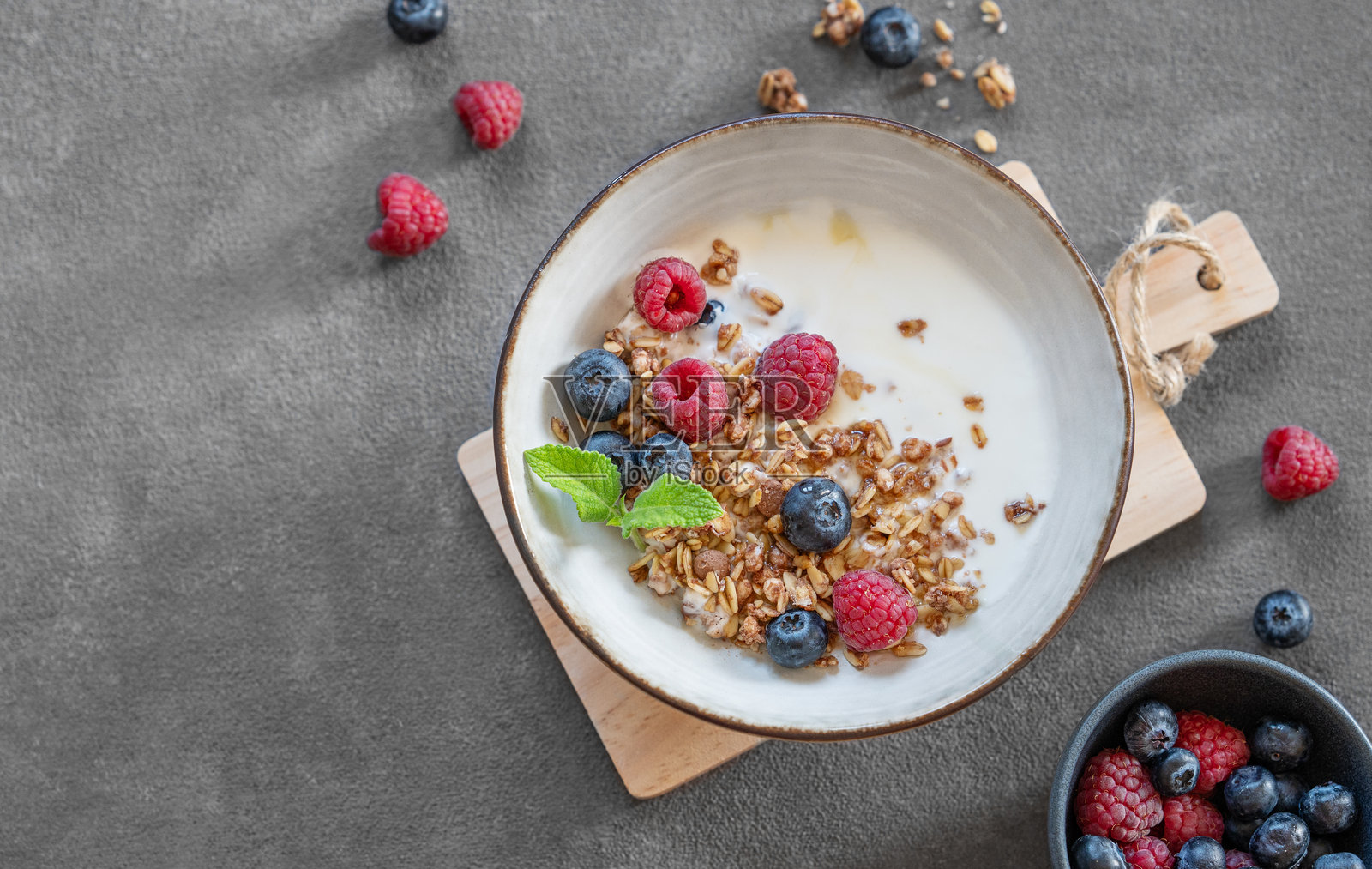 碗中盛有格兰诺拉麦片、希腊酸奶、覆盆子、蓝莓和蜂蜜,放在木板上,背景为深色,旁边有新鲜的浆果。照片摄影图片