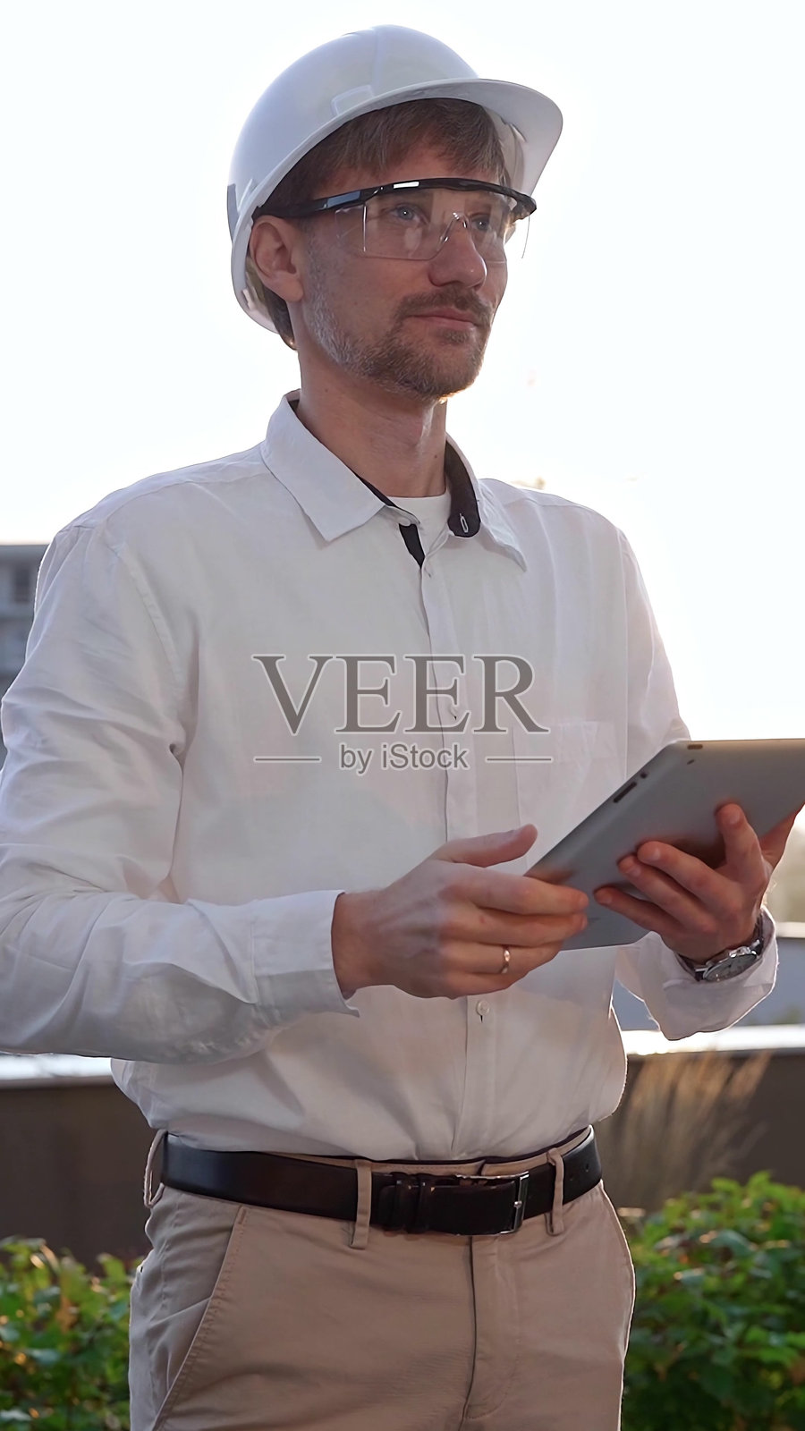 男性建筑工程师穿着白衬衫和安全帽,在日出时分使用数字平板电脑检查建筑工地,正面和垂直视角。建筑与工程照片摄影图片