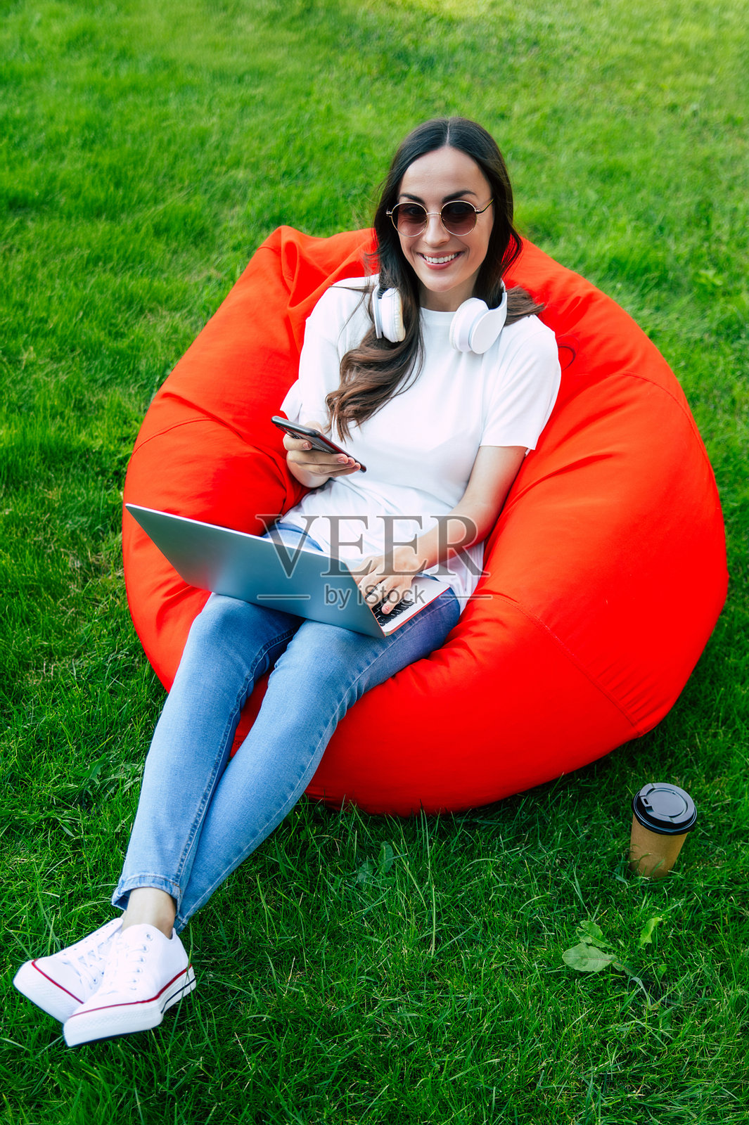 现代微笑自信的年轻女子的全身形象,穿着休闲服,戴着太阳镜,坐在绿色草坪花园的豆袋椅上,拿着笔记本电脑和手机。女性自由职业者,商业,工作照片摄影图片